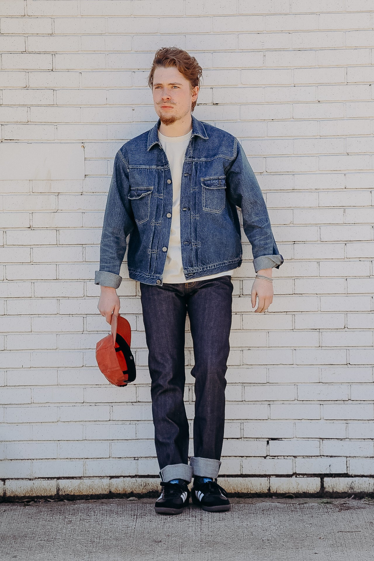 Man wearing denim jacket, white shirt, dark jeans, and black sneakers holding a red cap against white brick wall