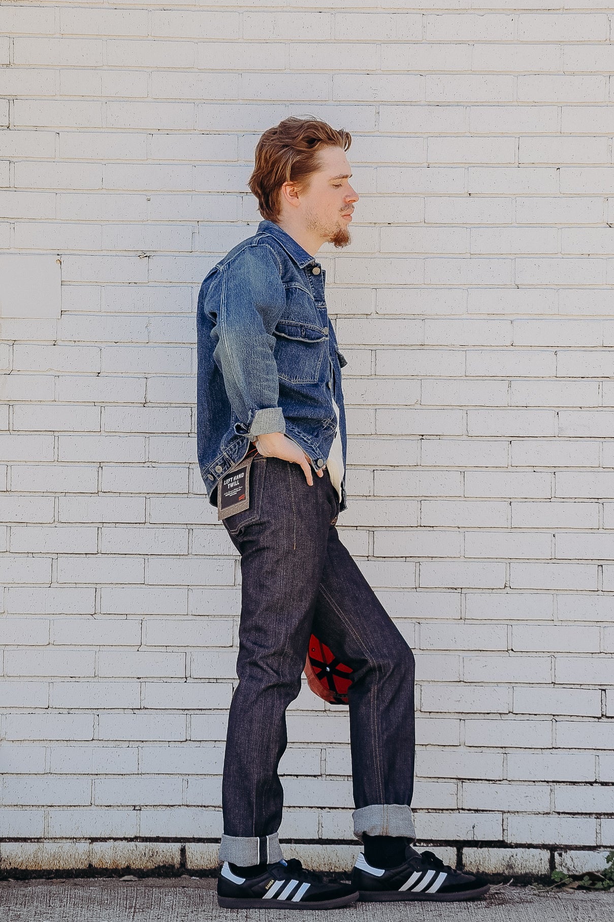 man wearing cuffed dark denim jeans and blue denim jacket standing by white brick wall holding red cap