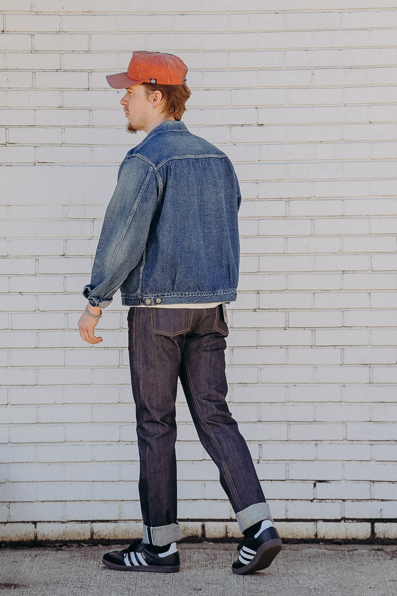 Man in blue denim jacket, dark jeans, and orange cap walking against white brick wall