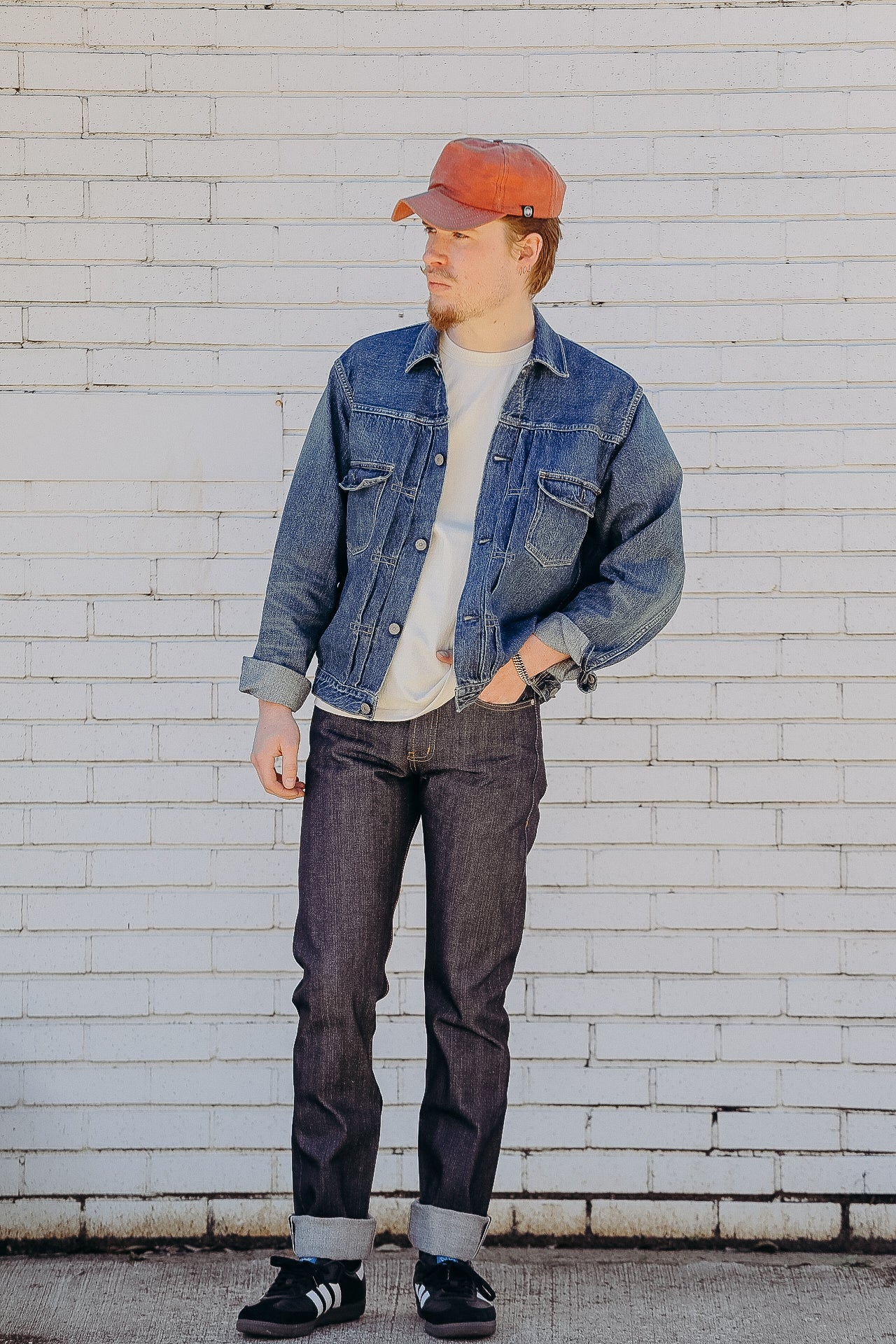 Man wearing denim jacket, dark jeans, white t-shirt, orange cap, and black sneakers standing against a white brick wall