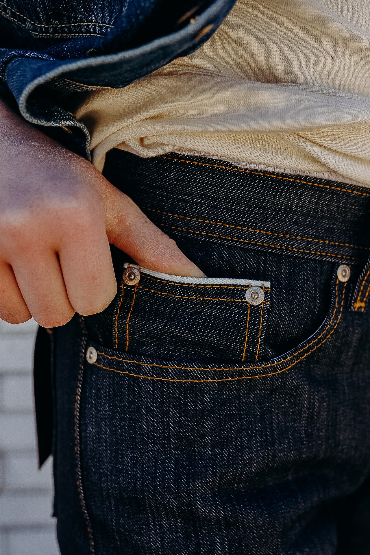 Close-up of hand reaching into small pocket of dark denim jeans with orange stitching, wearing beige shirt