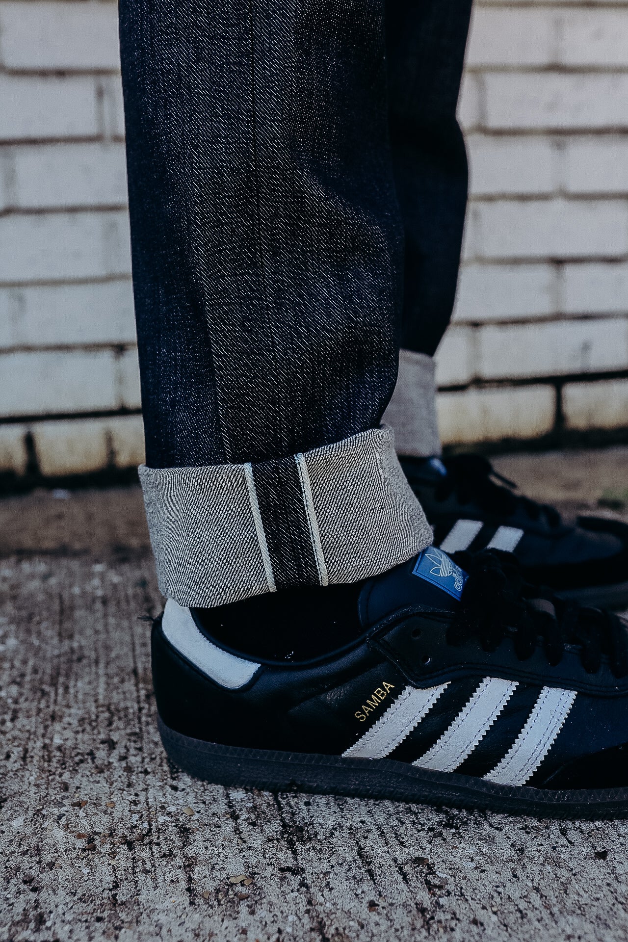 Close-up of rolled raw denim jeans paired with black Adidas Samba sneakers on textured concrete floor