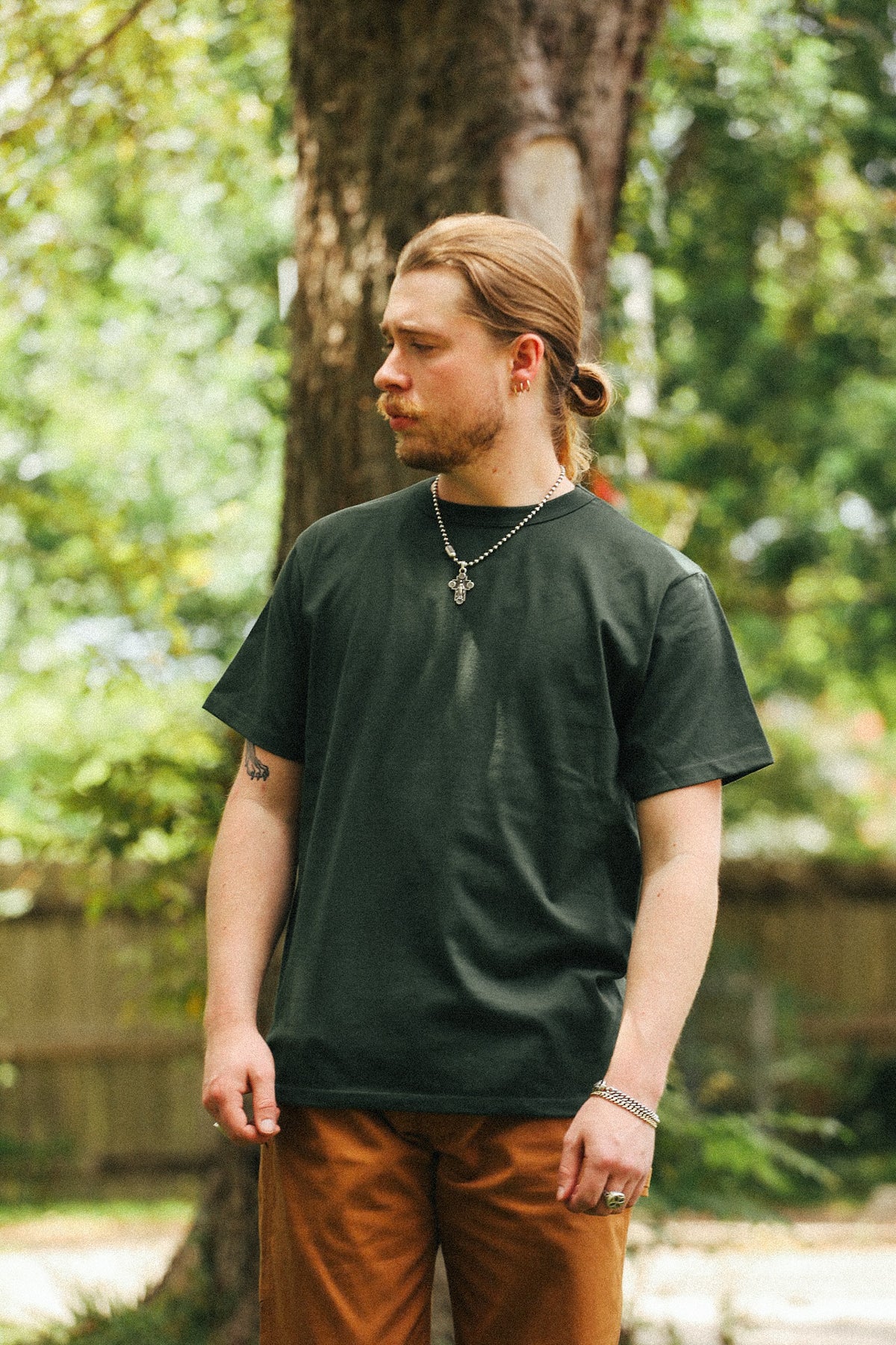 A man with blonde hair tied back stands outdoors in front of a tree, wearing a dark forest green short-sleeve t-shirt and brown pants.