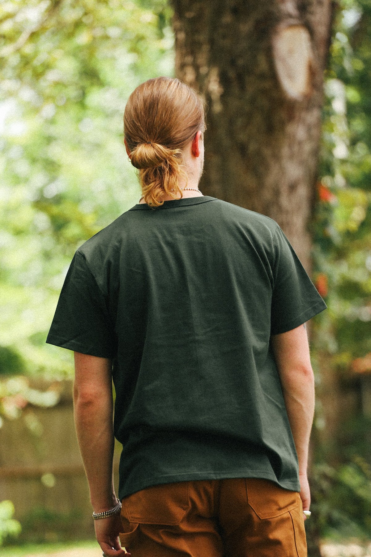 A man with blonde hair tied back stands outdoors in front of a tree, wearing a dark forest green short-sleeve t-shirt and brown pants.