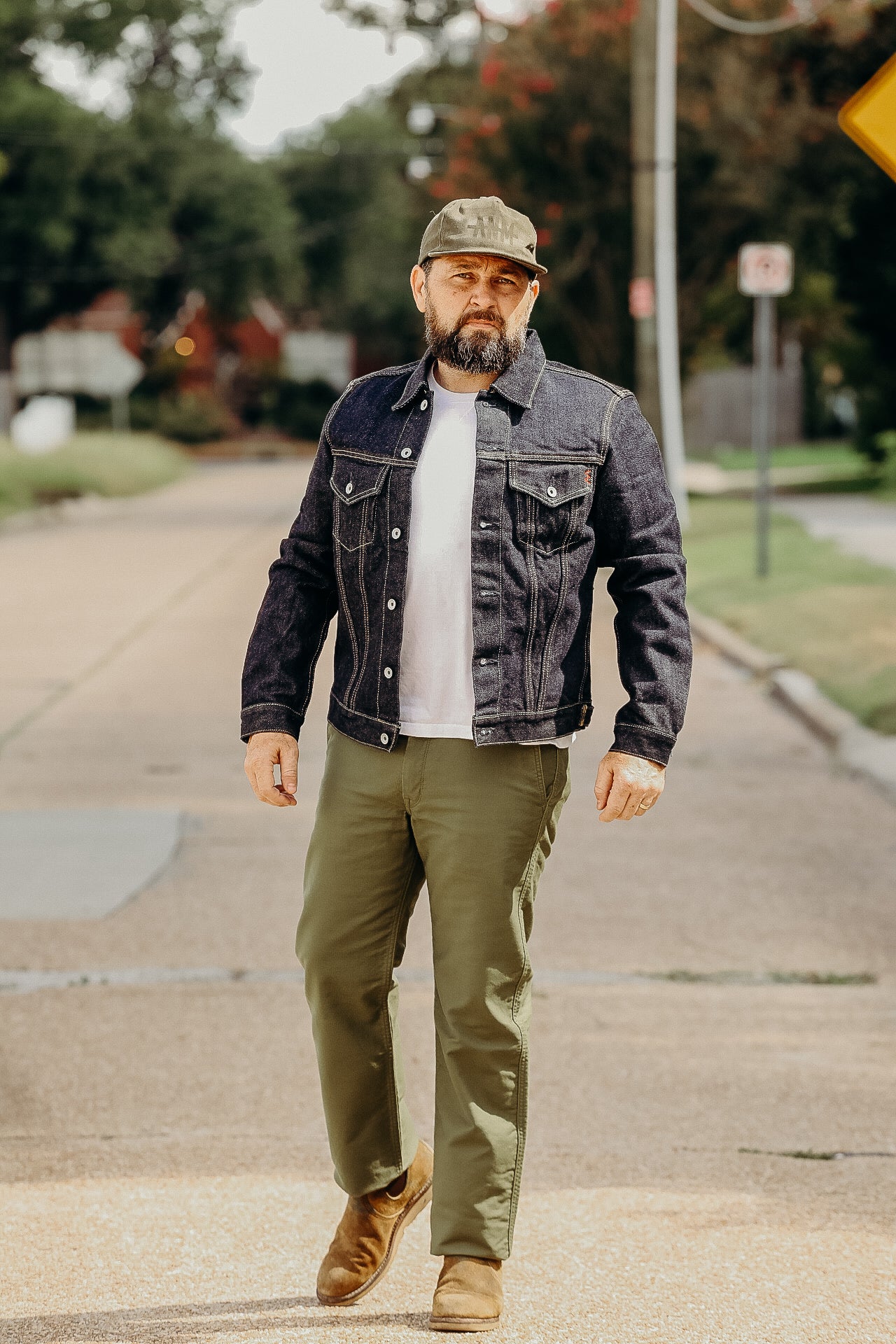 Man wearing dark denim jacket, white t-shirt, olive pants, tan boots, and green cap walking outdoors on street