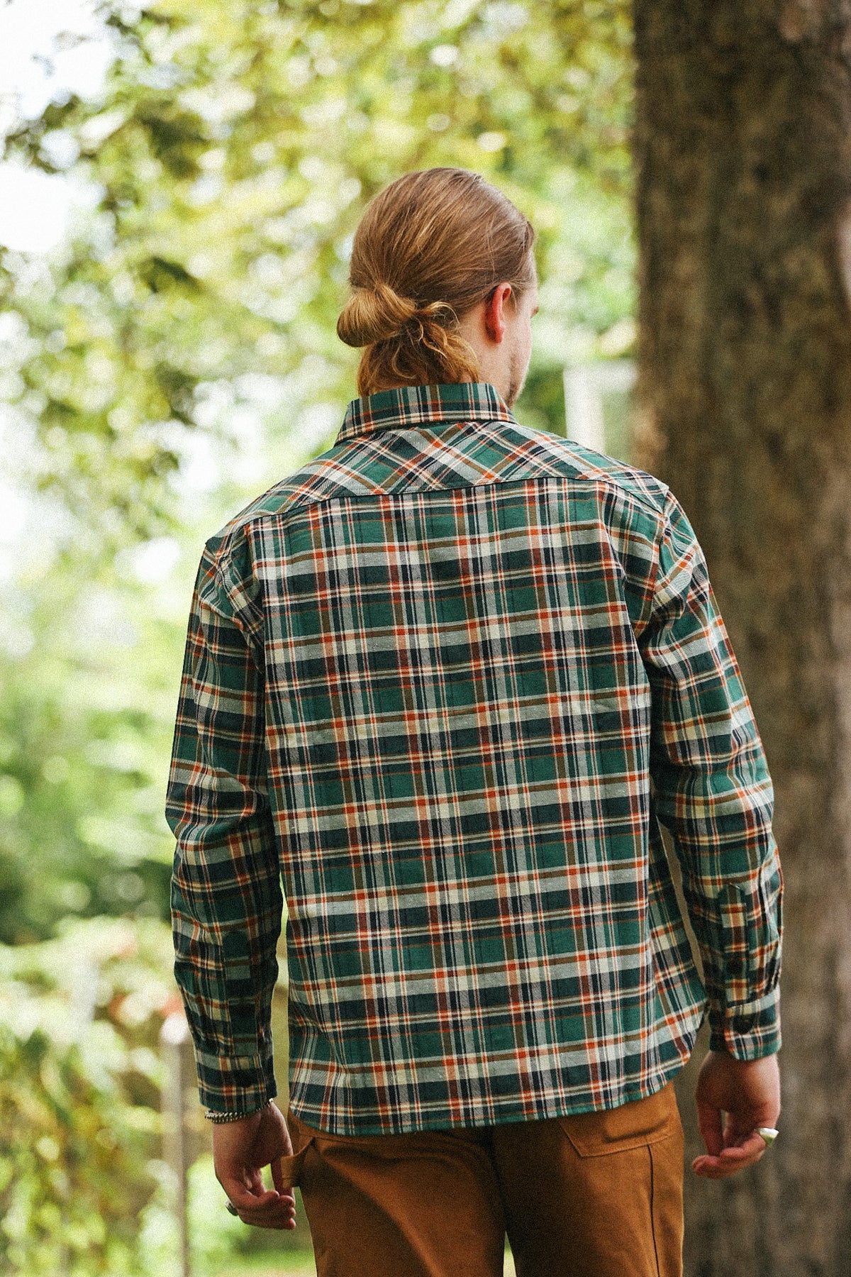 A man viewed from behind wearing a green plaid flannel shirt with brown pants, standing outdoors in front of a tree.