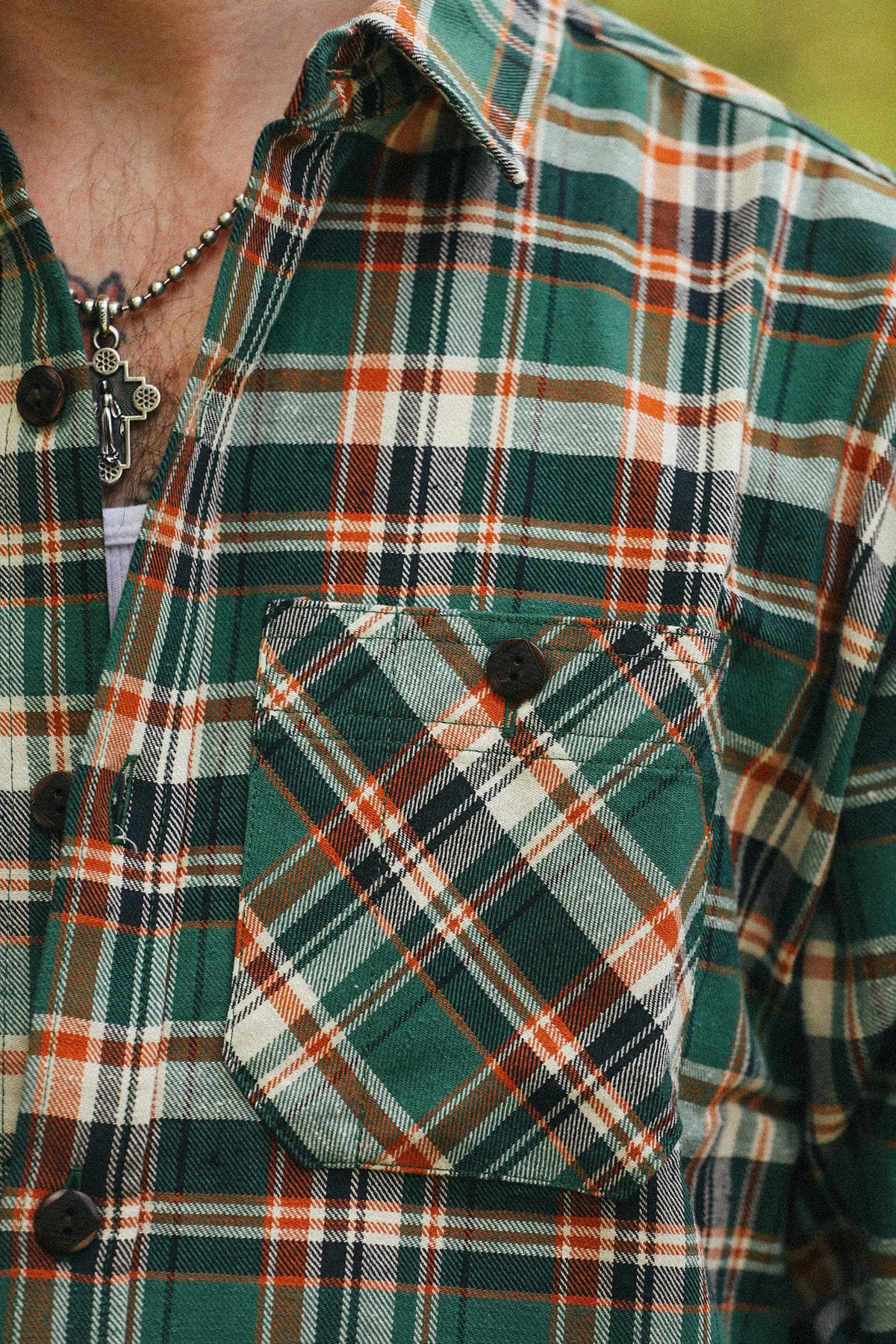 Close-up of a green plaid flannel shirt with orange, white, and black stripes, featuring a chest pocket and dark buttons.