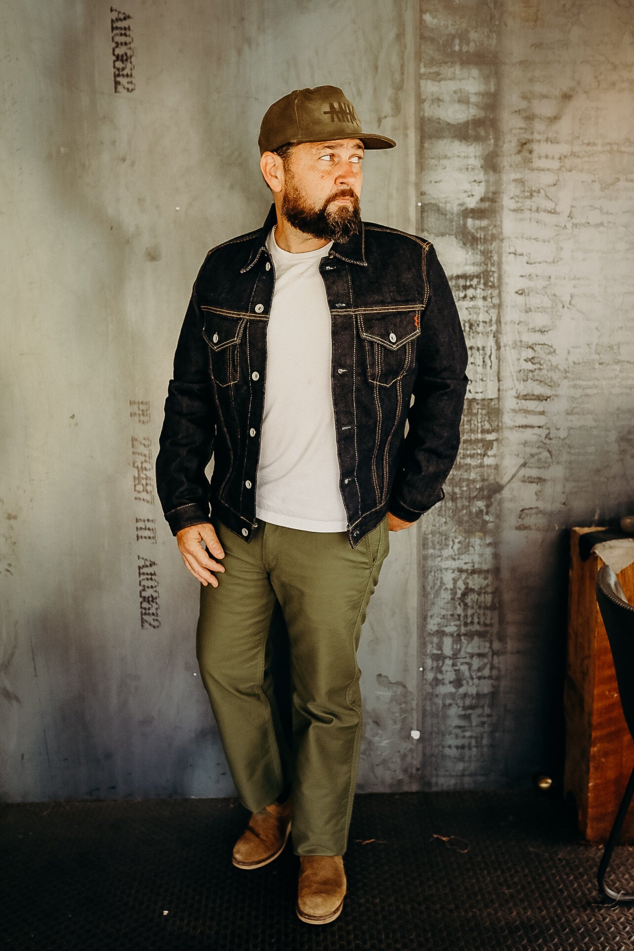 Bearded man wearing black denim jacket, white shirt, olive pants, and brown boots standing indoors