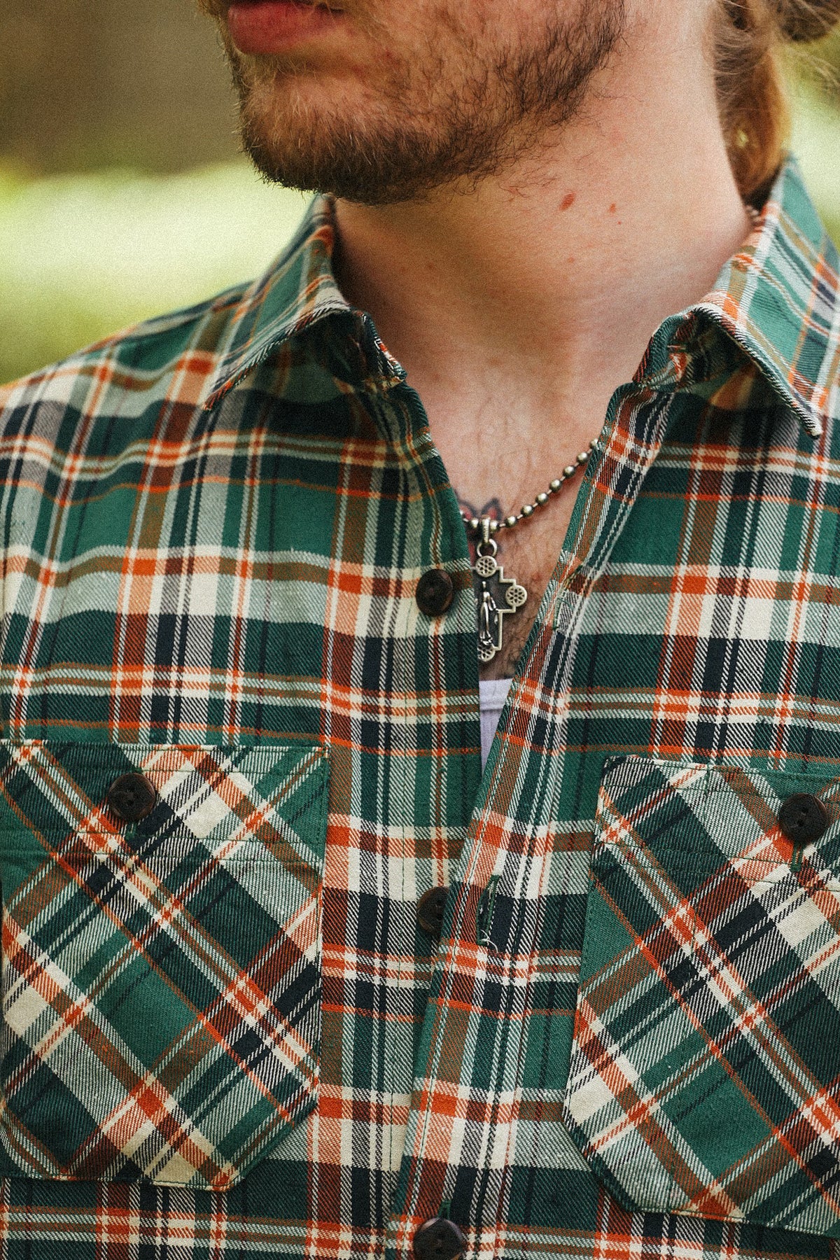 A close-up of a man wearing a green, orange, and white plaid flannel shirt, featuring a silver cross pendant necklace.