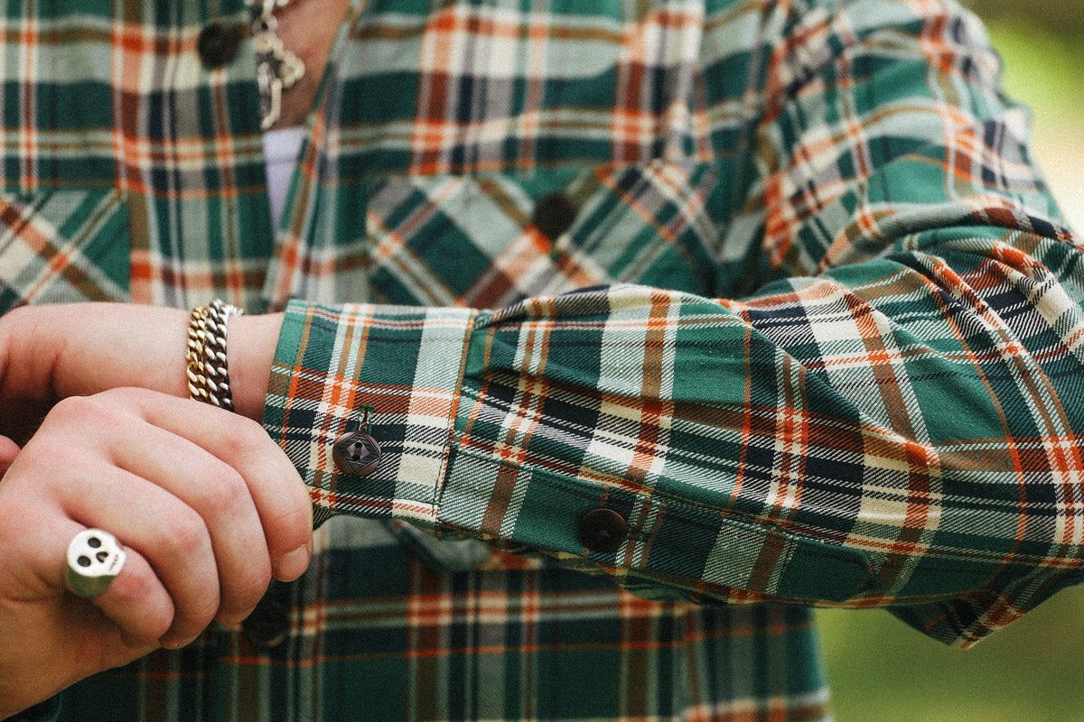 Close-up of a person wearing a green, orange, and white plaid flannel shirt, buttoning the cuff with a silver-toned ring on one finger.