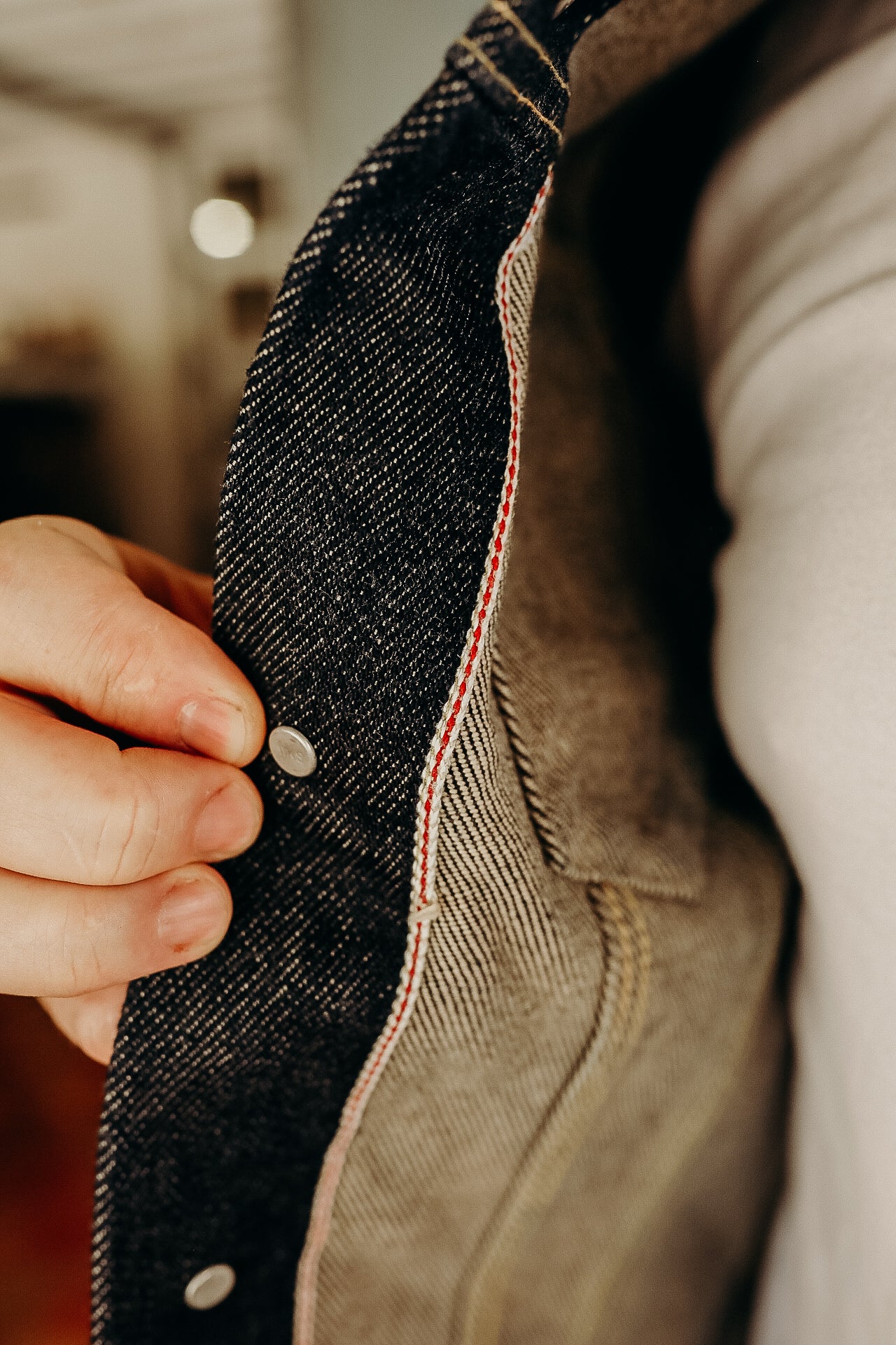 Close-up of hand holding open raw selvedge denim jacket showing red stitch detail and metal snap button