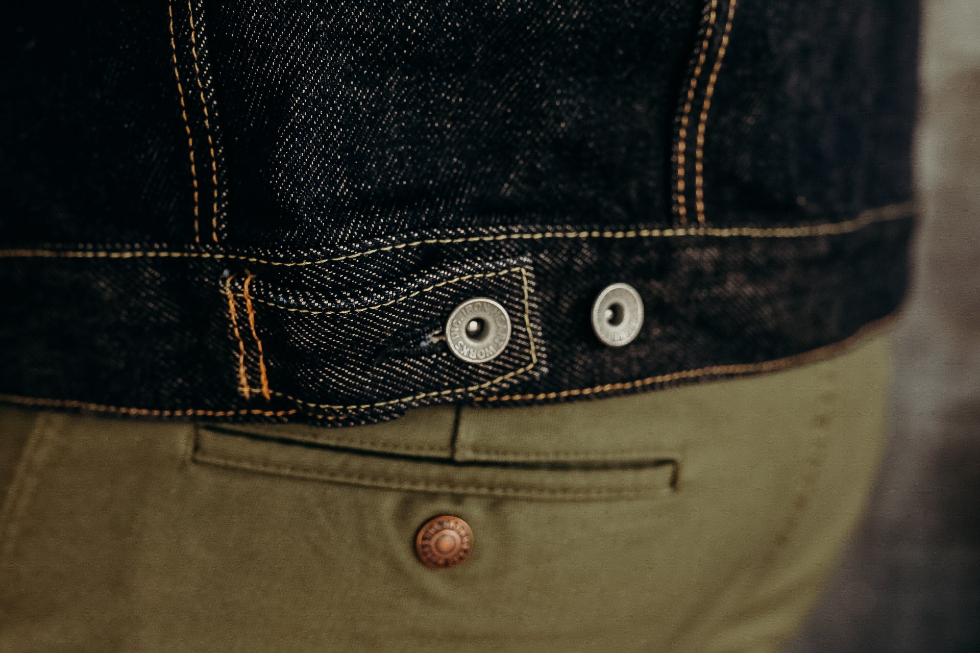 Close-up of dark denim jacket with contrast stitching and metal buttons paired with olive green pants at Iron Shop Provisions