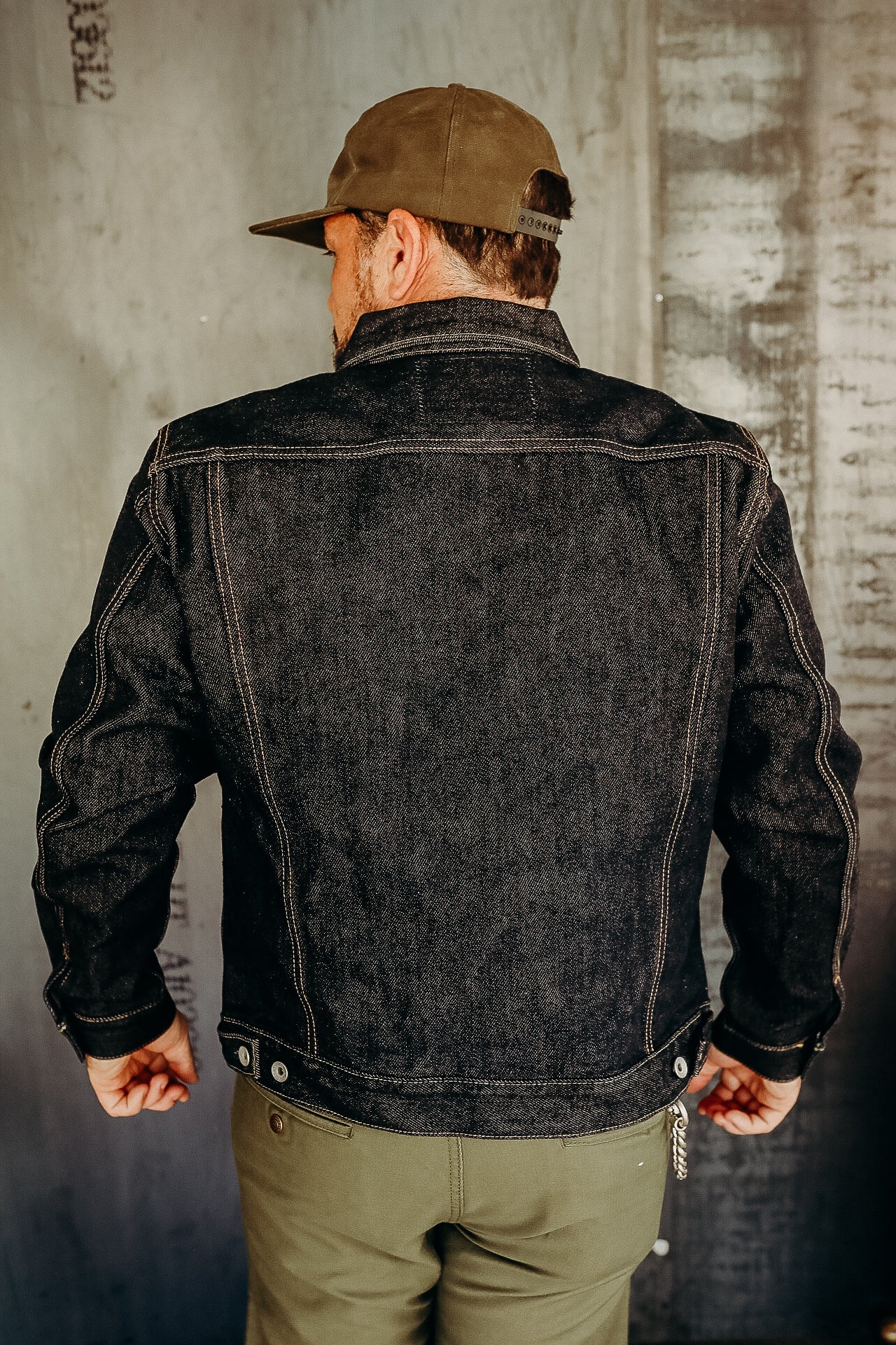 Man wearing a dark denim jacket and olive green pants with cap standing against distressed concrete wall