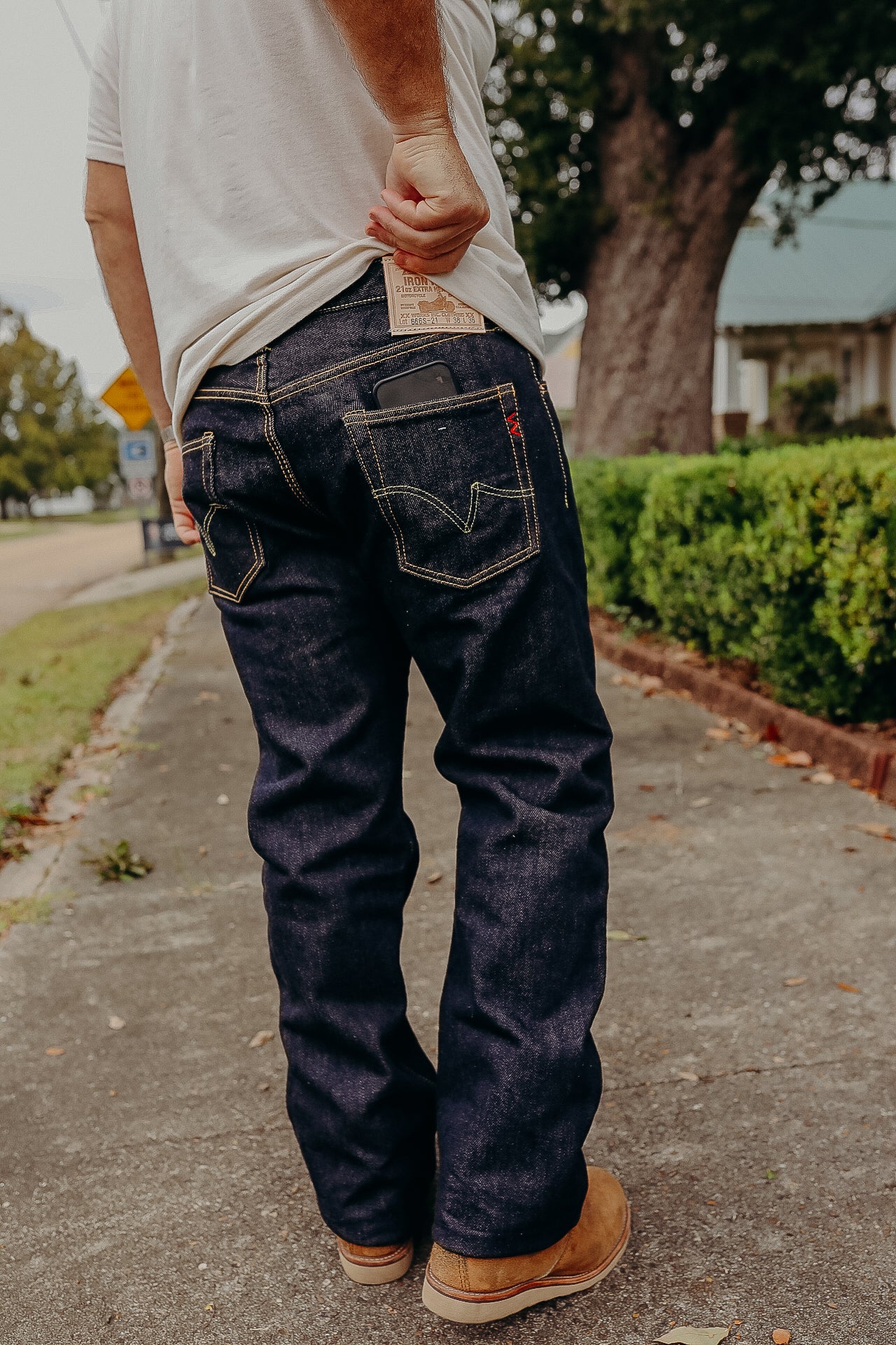 Person wearing dark raw denim jeans with a phone in back pocket, standing on sidewalk near bushes
