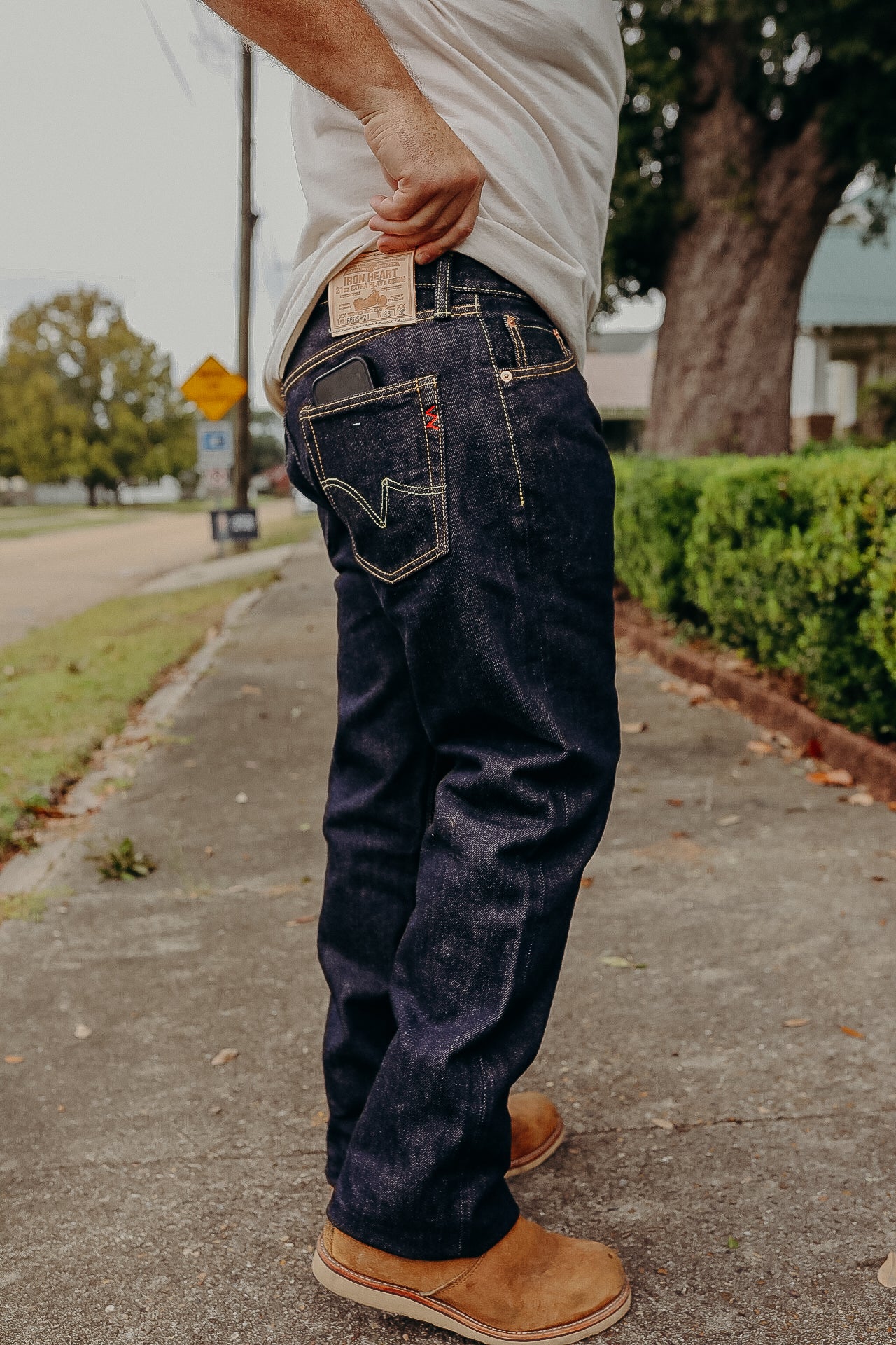 Person wearing dark raw denim jeans and tan work boots outdoors on sidewalk, holding jeans label