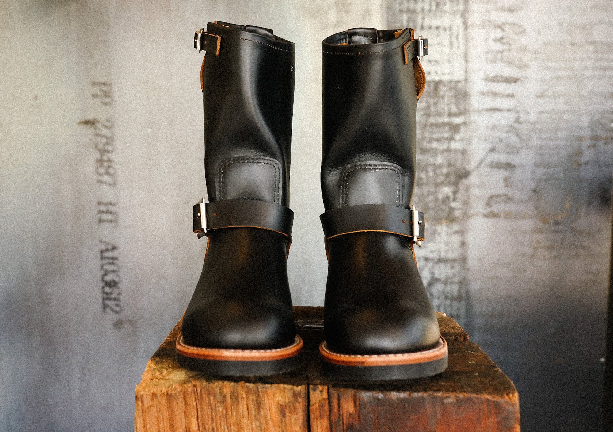 Pair of black leather engineer boots with buckles on rustic wooden block against textured wall
