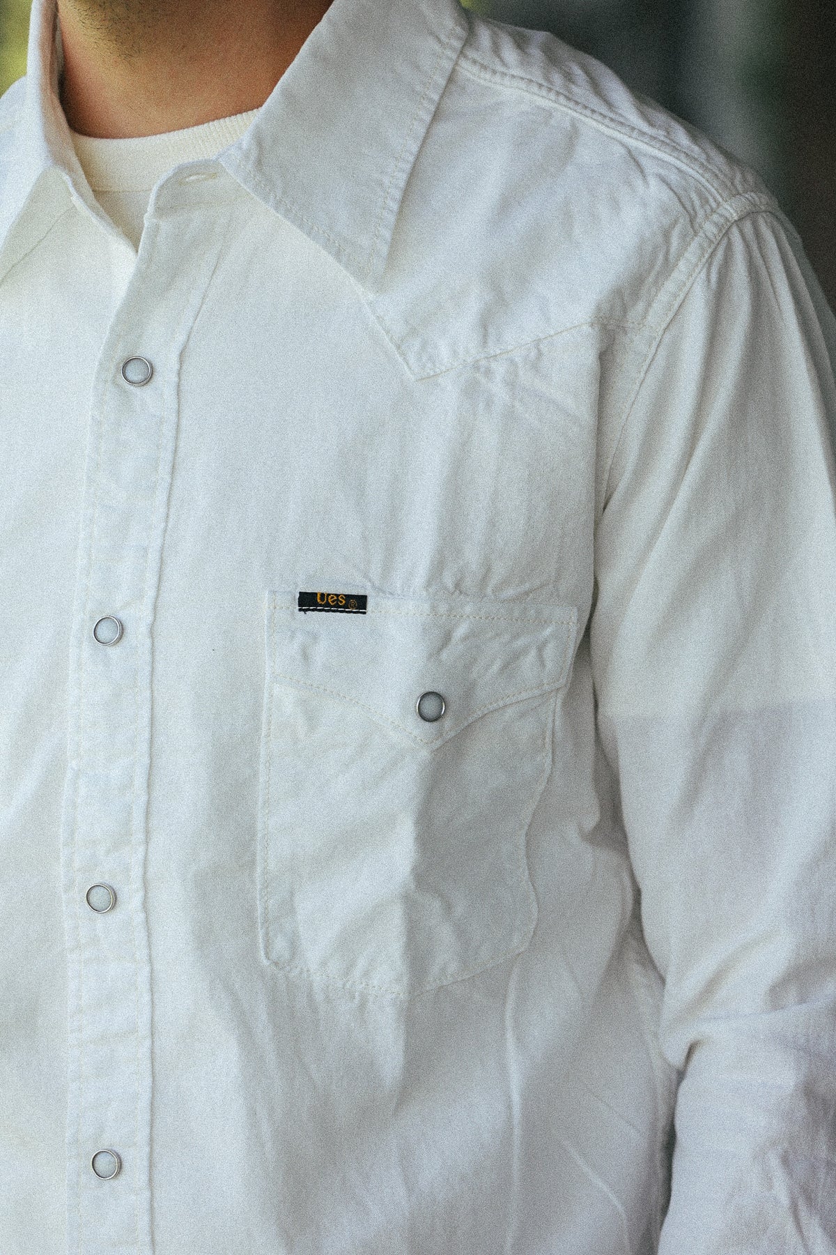 A close-up of a person wearing a white long-sleeve button-up shirt with a chest pocket and a small black brand label.
