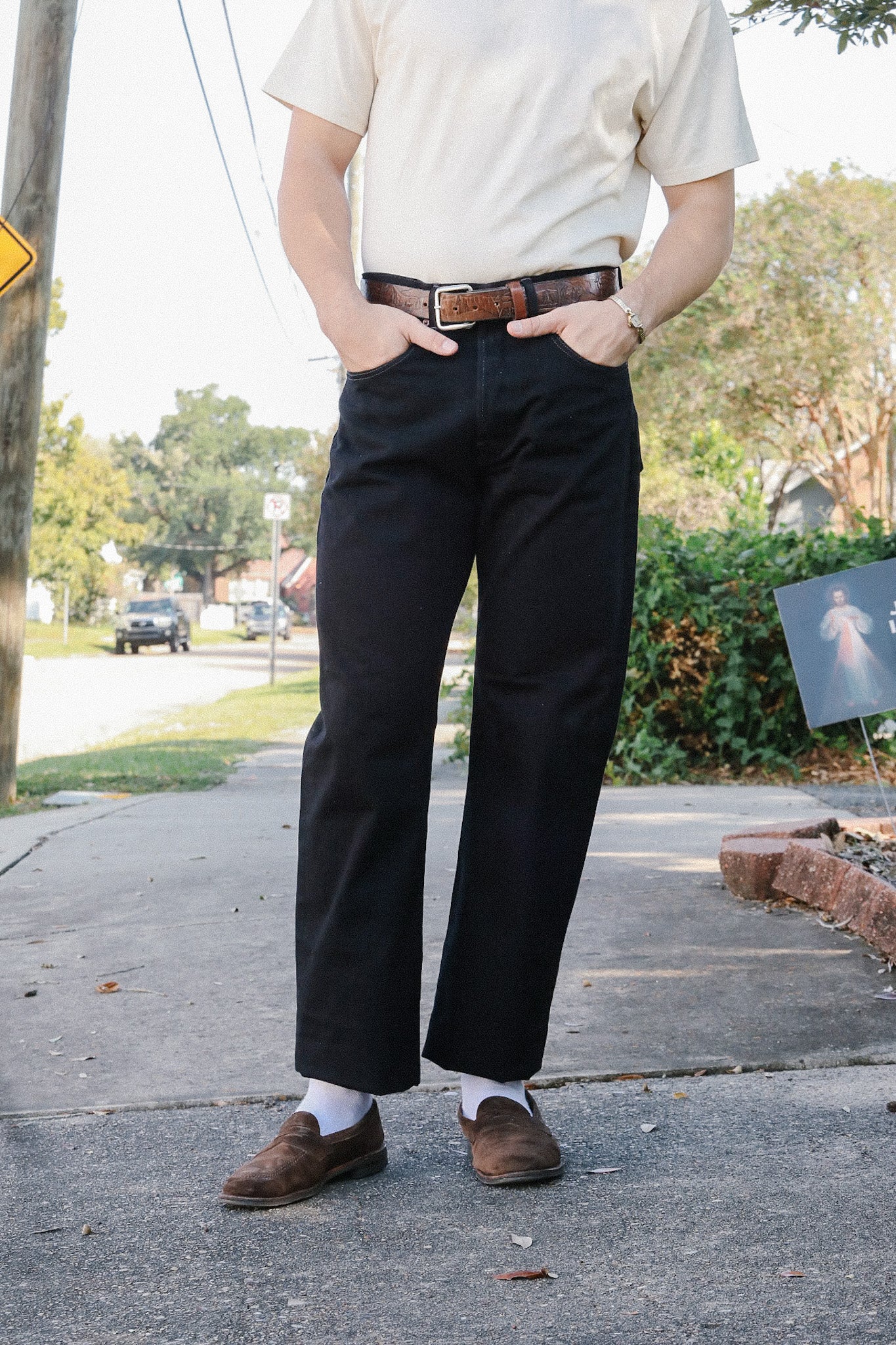 Person wearing black pants with brown leather belt and brown loafers, hands in pockets on sidewalk