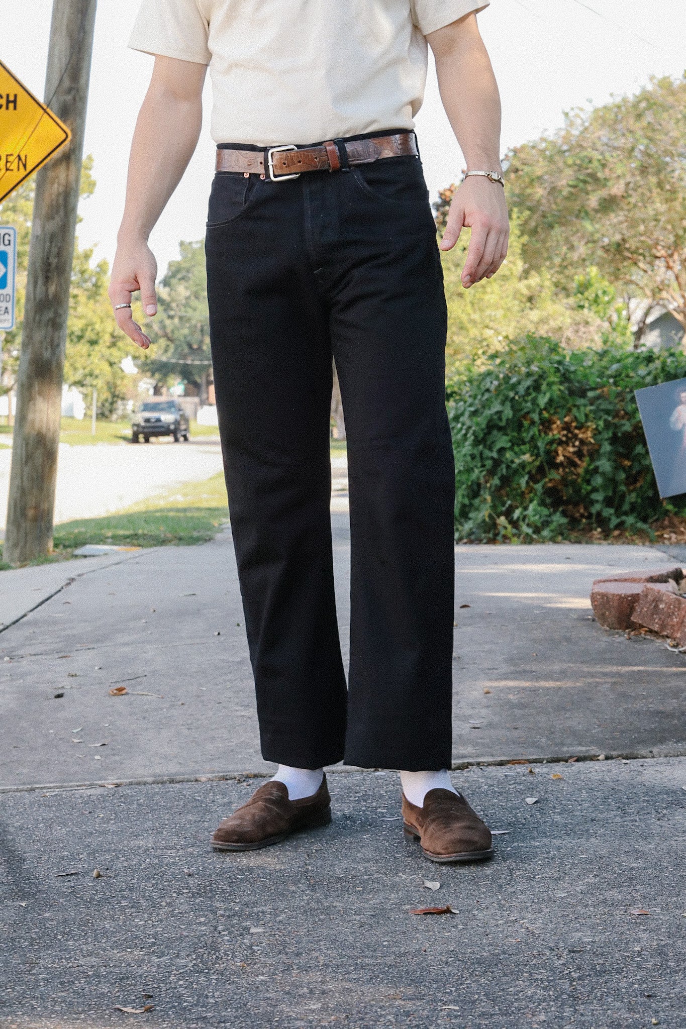 Man wearing black pants, brown leather belt, white socks, and brown suede shoes standing on sidewalk