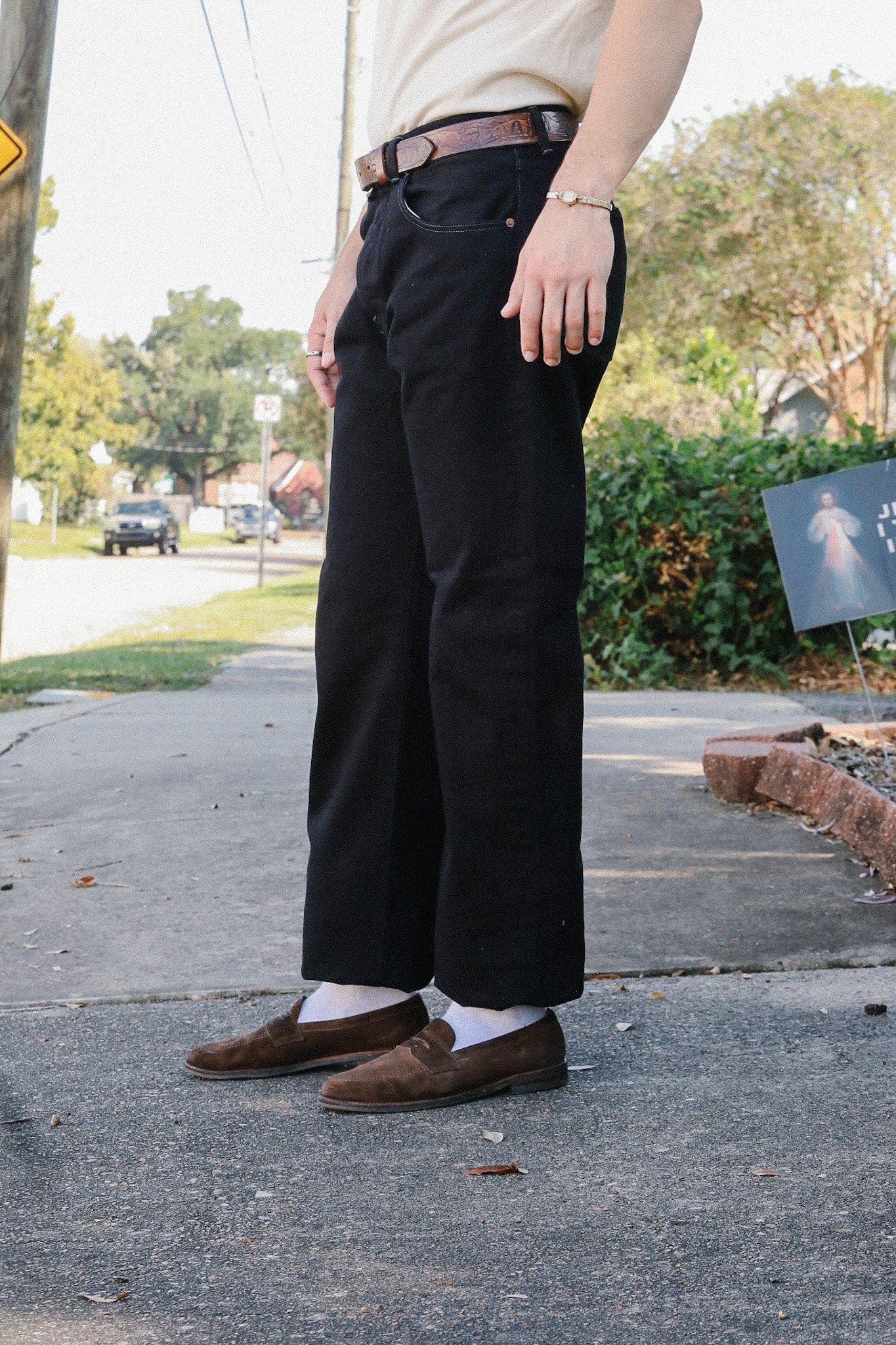 Person wearing black casual pants, brown belt, brown loafers, and white socks standing outdoors on a sidewalk