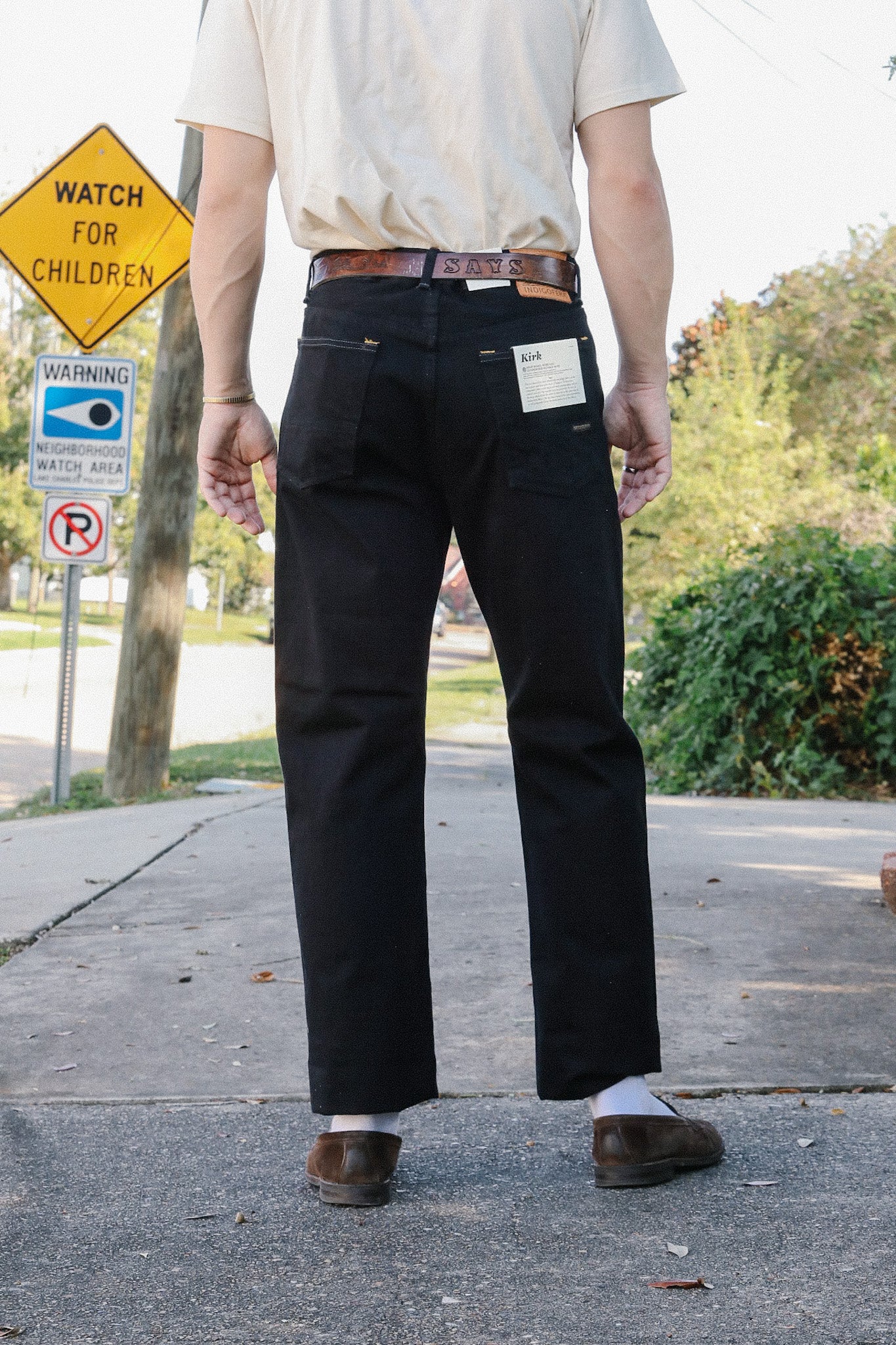 Man wearing black Kirk jeans and brown leather belt standing on sidewalk near watch for children street sign