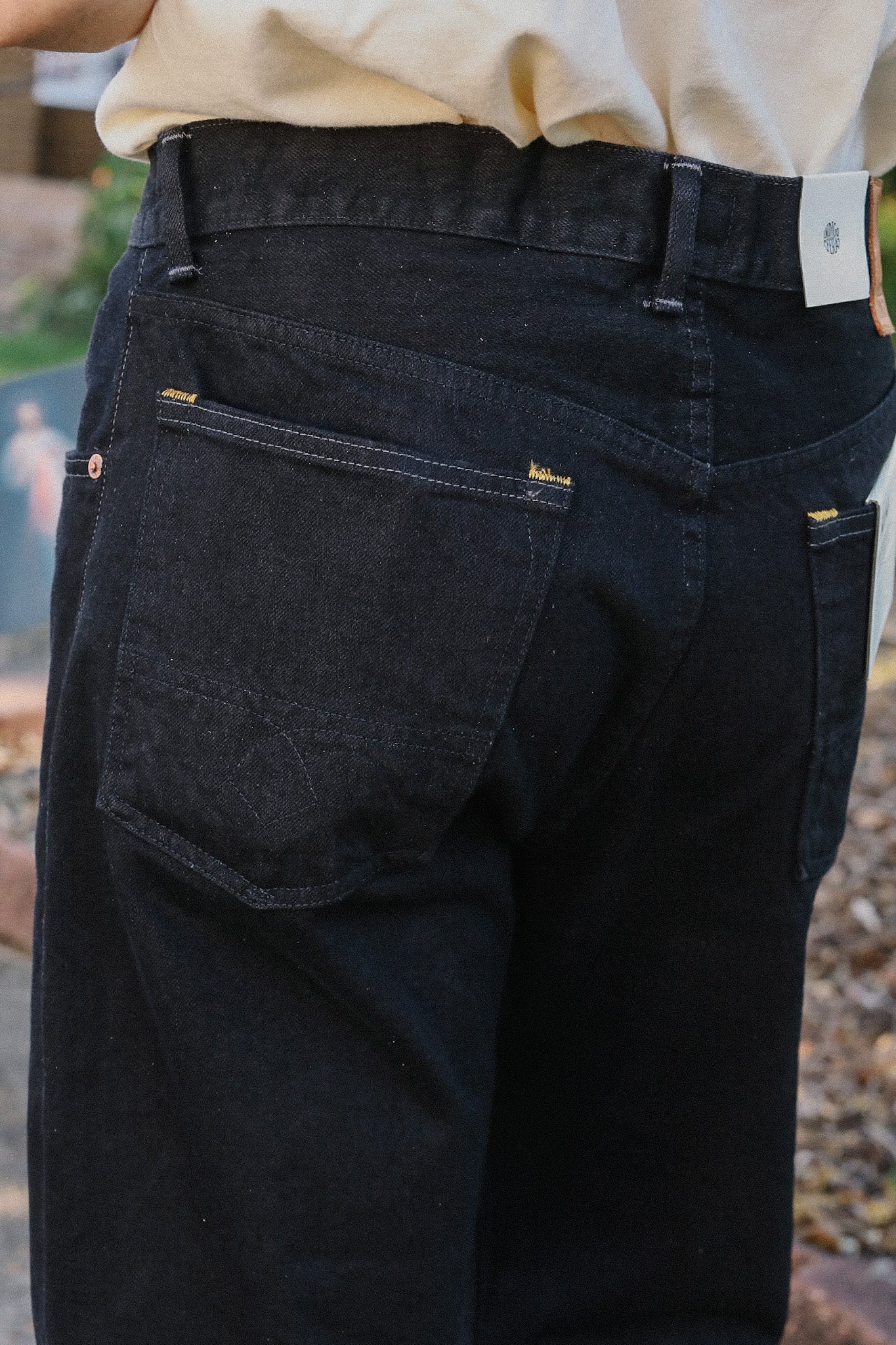 Close-up of black jeans with visible stitching and a fabric tag, worn with a light shirt outdoors