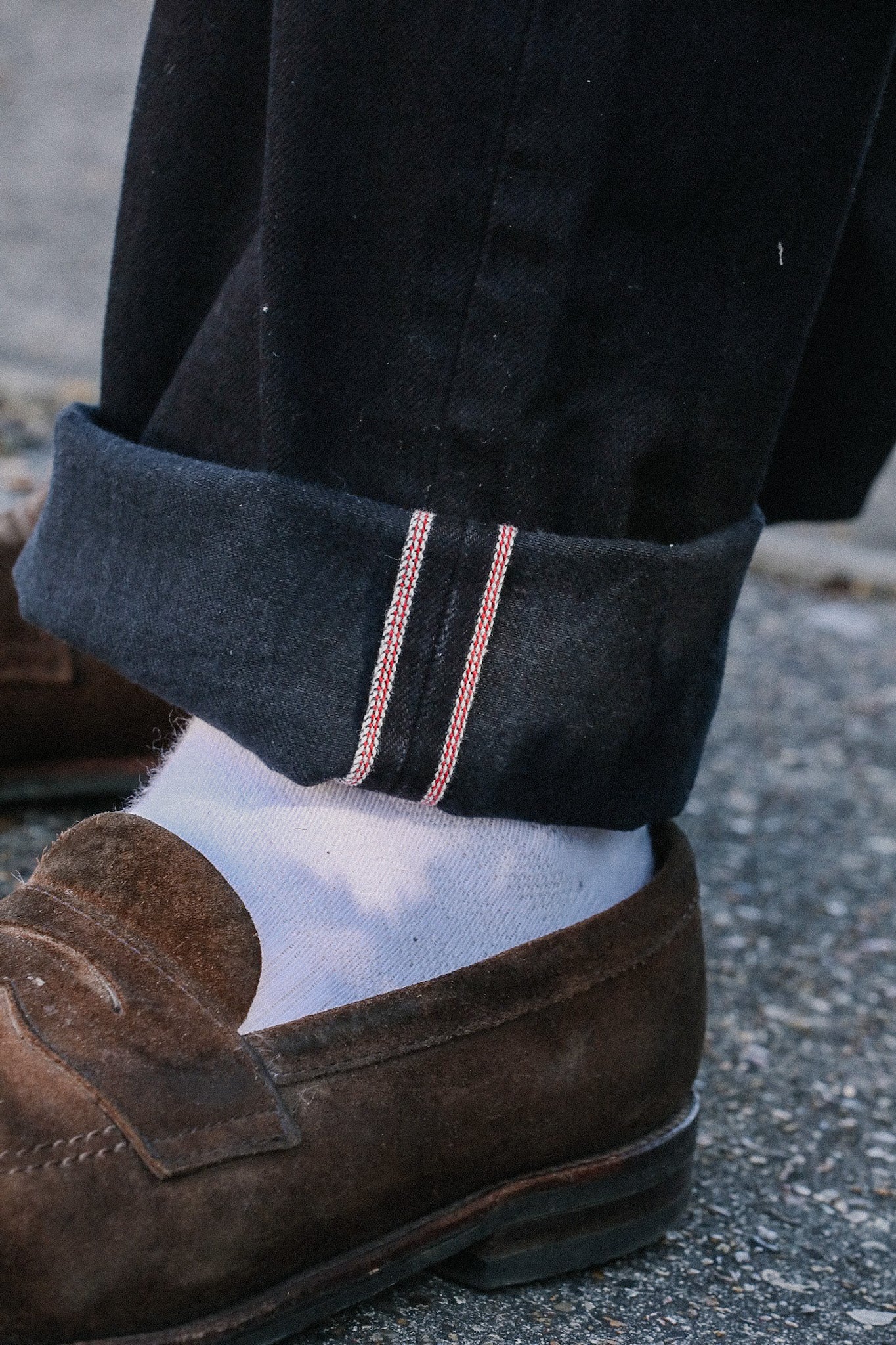 Close-up of rolled black denim jeans with red and white selvedge edge, white socks, and brown suede loafers on pavement