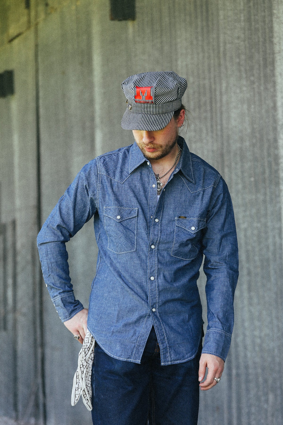 A man wearing a blue denim long-sleeve button-up shirt, dark jeans, a gray striped cap with a red logo patch, and holding a white knit sweater, standing against a textured gray concrete wall.