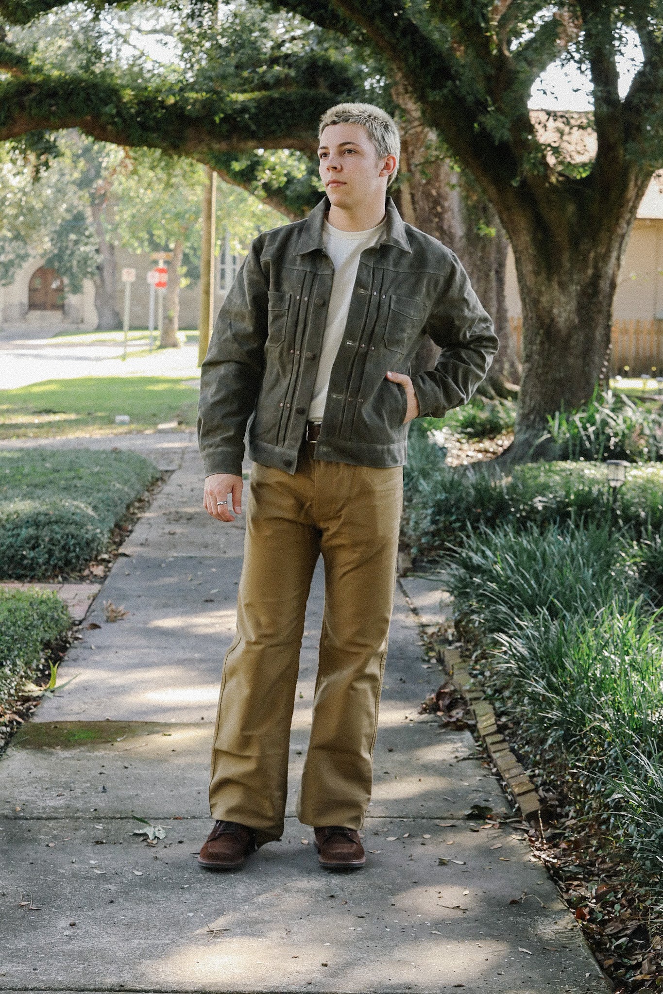 Young person wearing a dark green rugged jacket and khaki pants standing on a sidewalk under large trees in a sunny neighborhood
