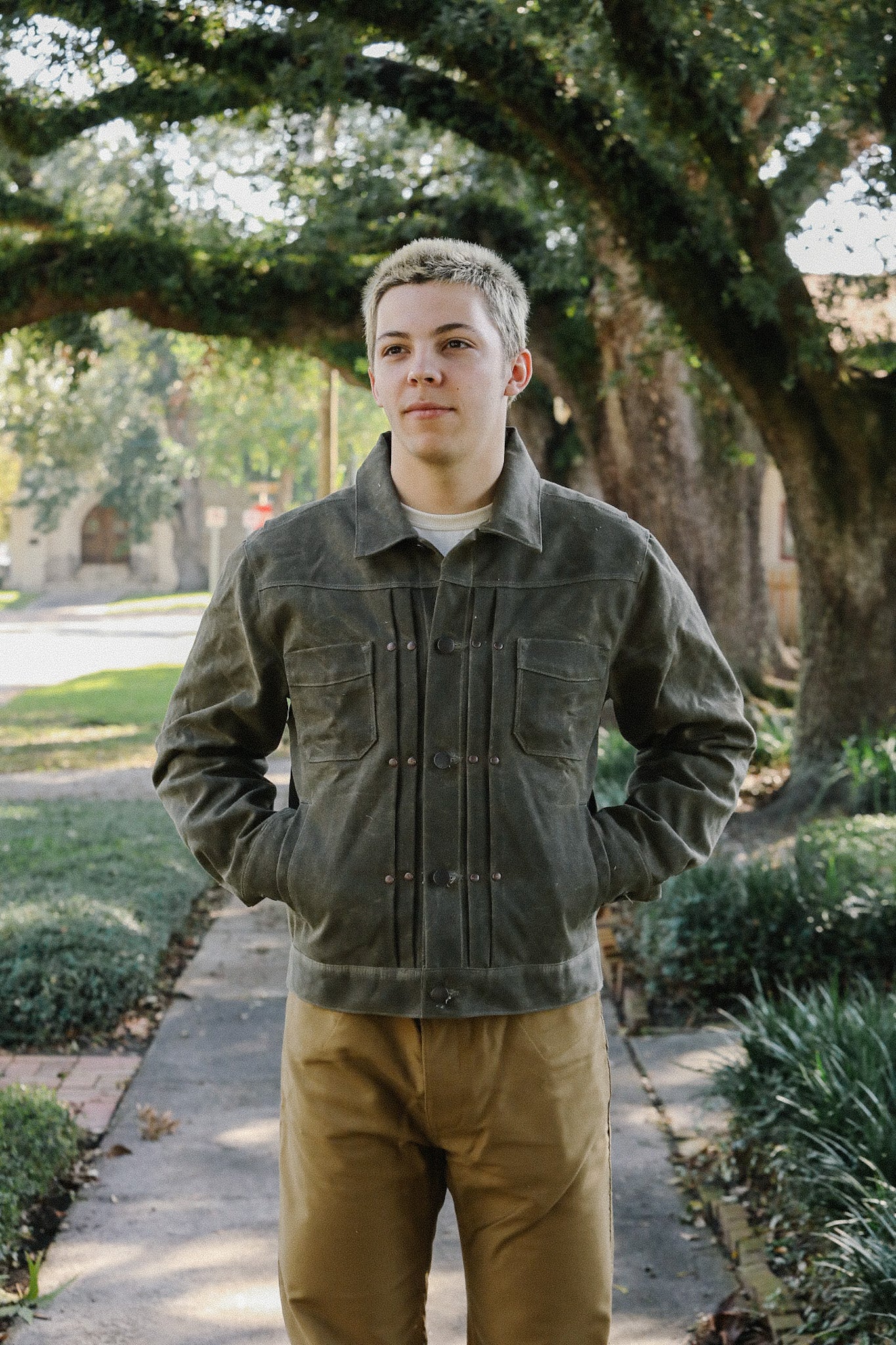 Young man wearing olive green rugged iron jacket and tan pants standing on a tree-lined sidewalk