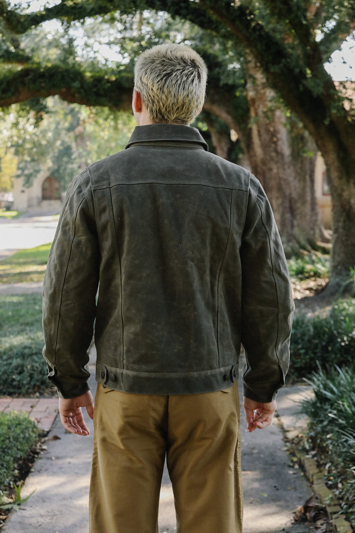 Man wearing olive green durable canvas jacket and brown pants standing on a tree-lined sidewalk outdoors