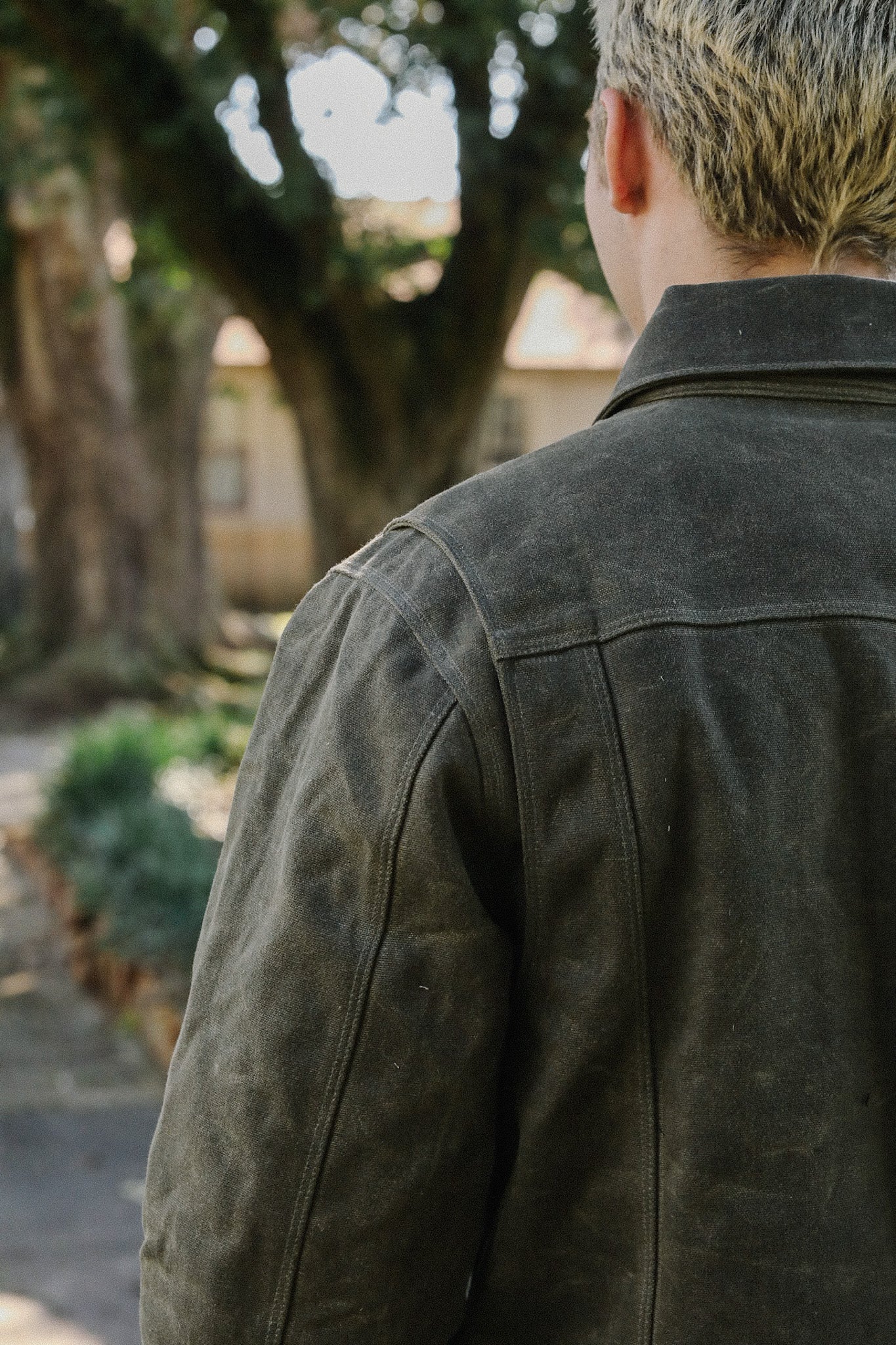 Back view of person wearing dark rugged jacket standing outdoors near trees and house