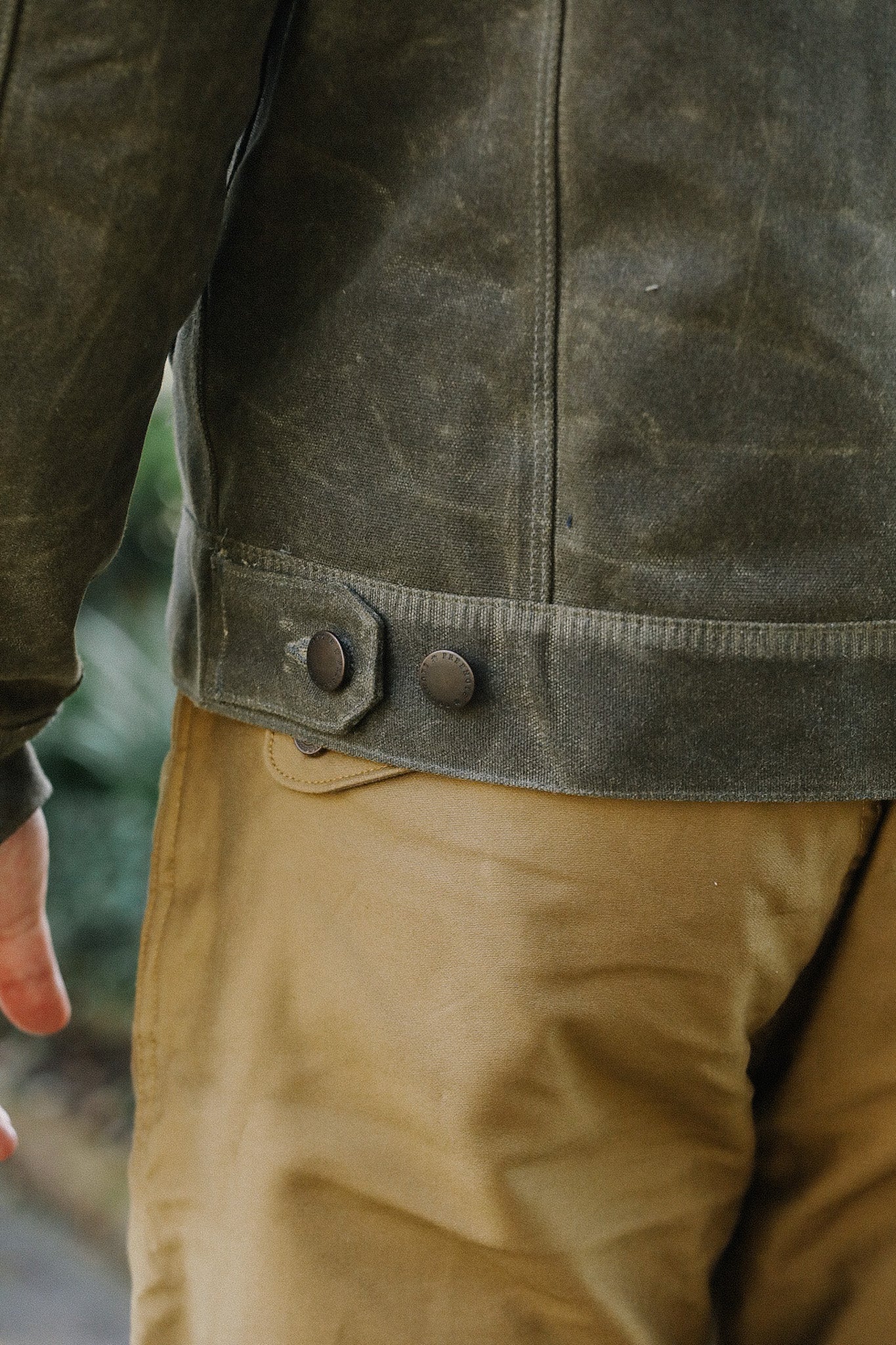 Close-up of a rugged olive canvas jacket with adjustable button strap paired with tan work pants