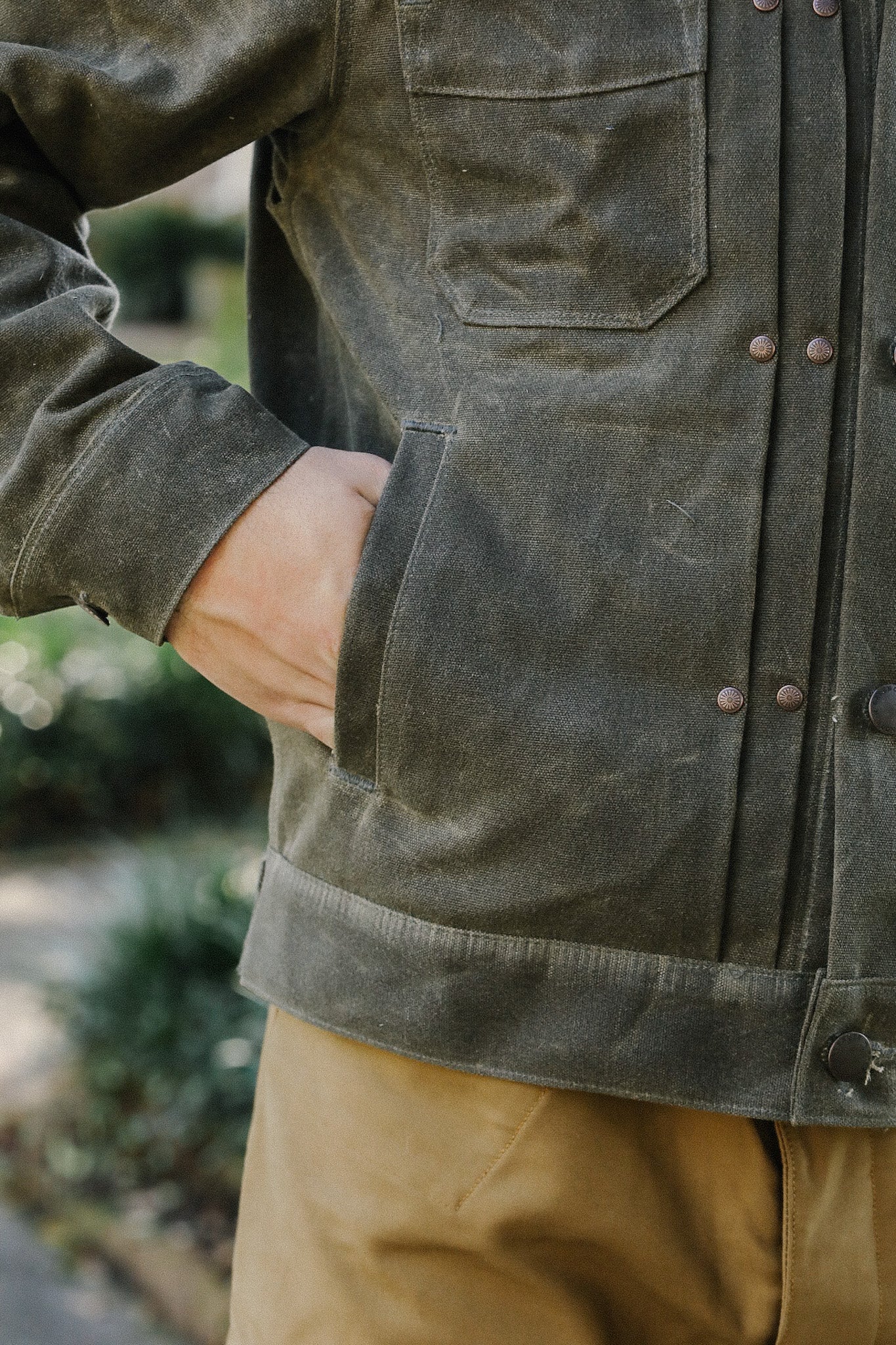Close-up of person wearing olive green work jacket with hand in pocket and tan pants outdoors