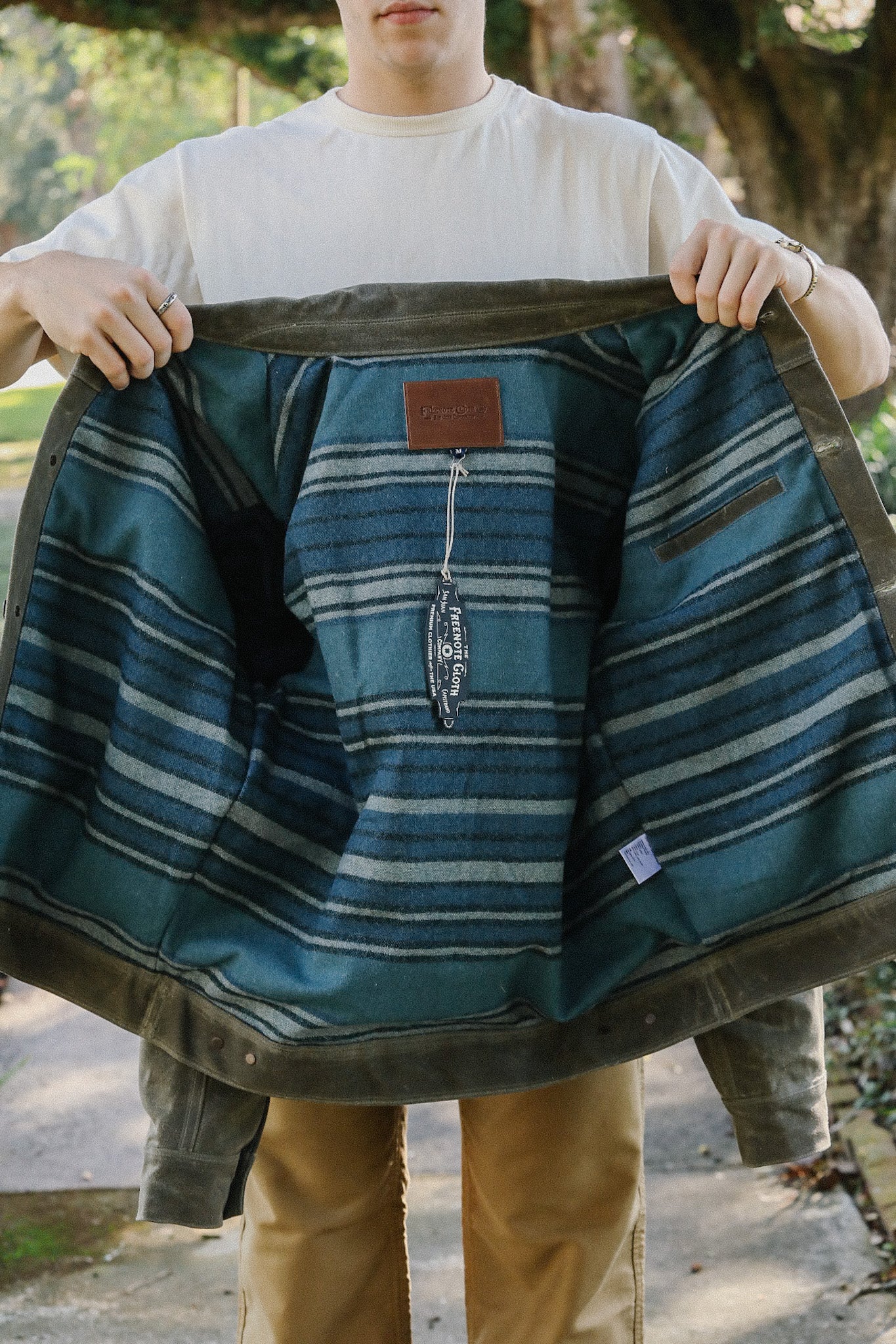Person holding open an olive green jacket revealing blue striped lining and tag in outdoor setting