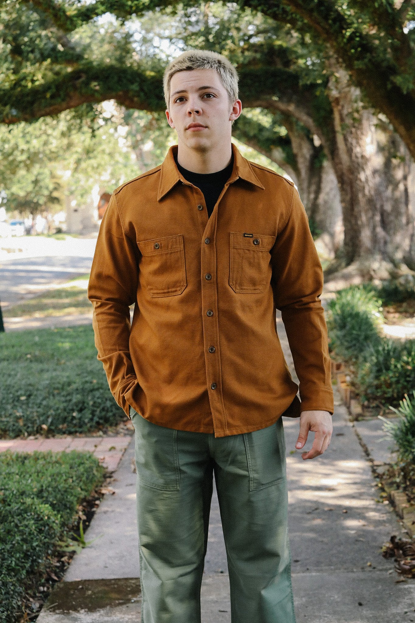 Man wearing brown work shirt and green utility pants standing on a tree-lined sidewalk