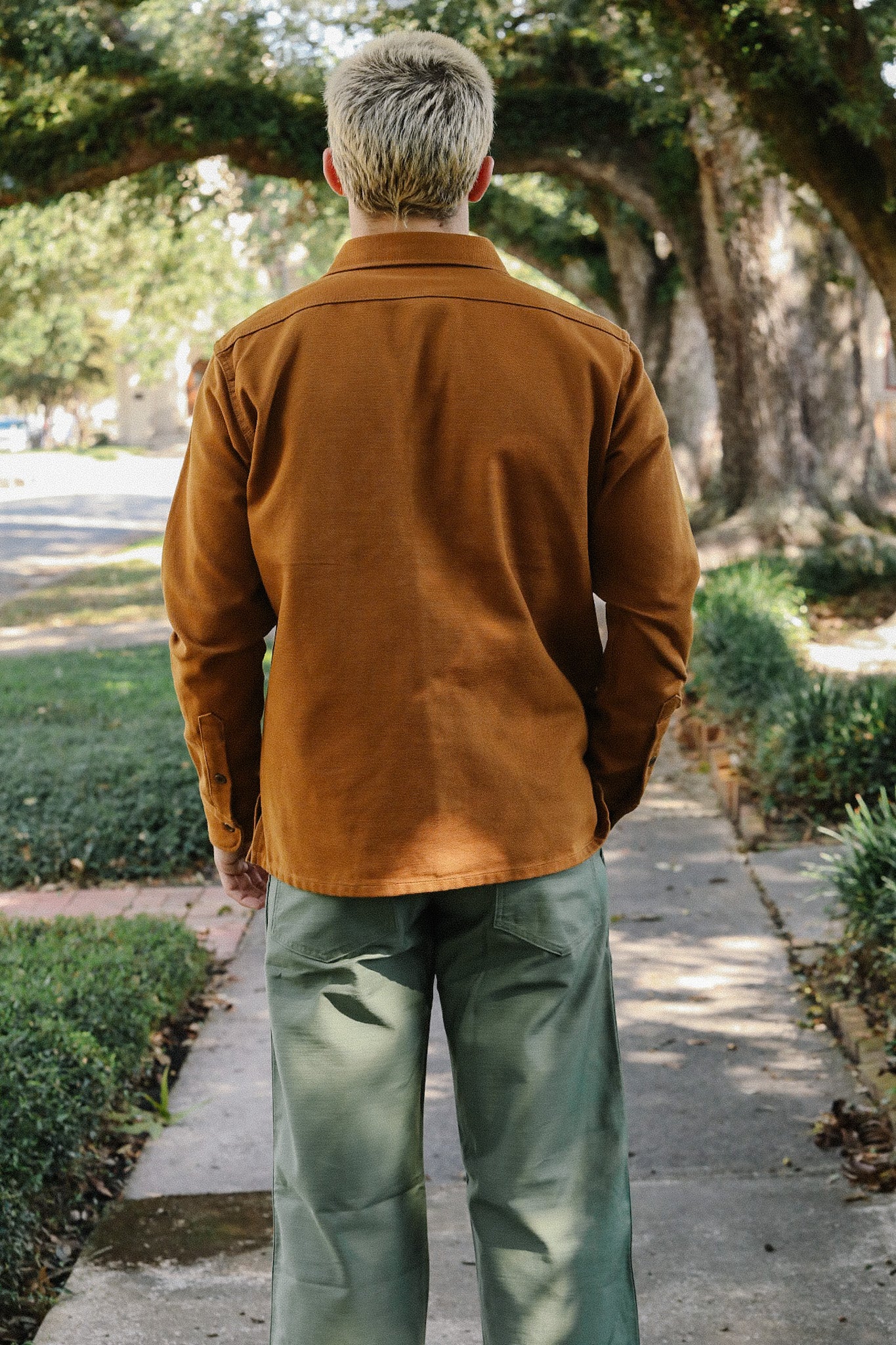 Man standing outdoors wearing brown shirt jacket and green pants on tree-lined sidewalk