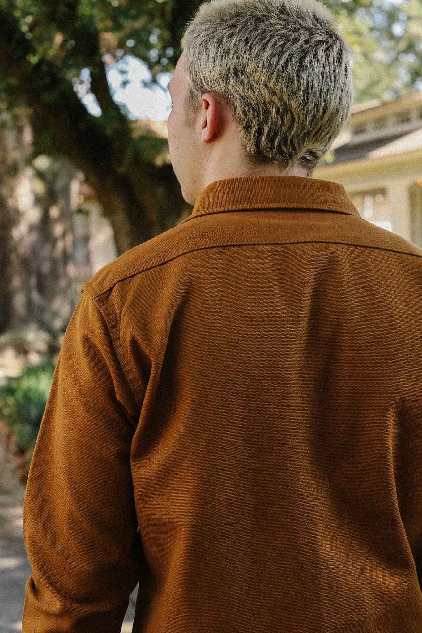 Person with short blonde hair wearing a brown jacket outdoors near trees and buildings