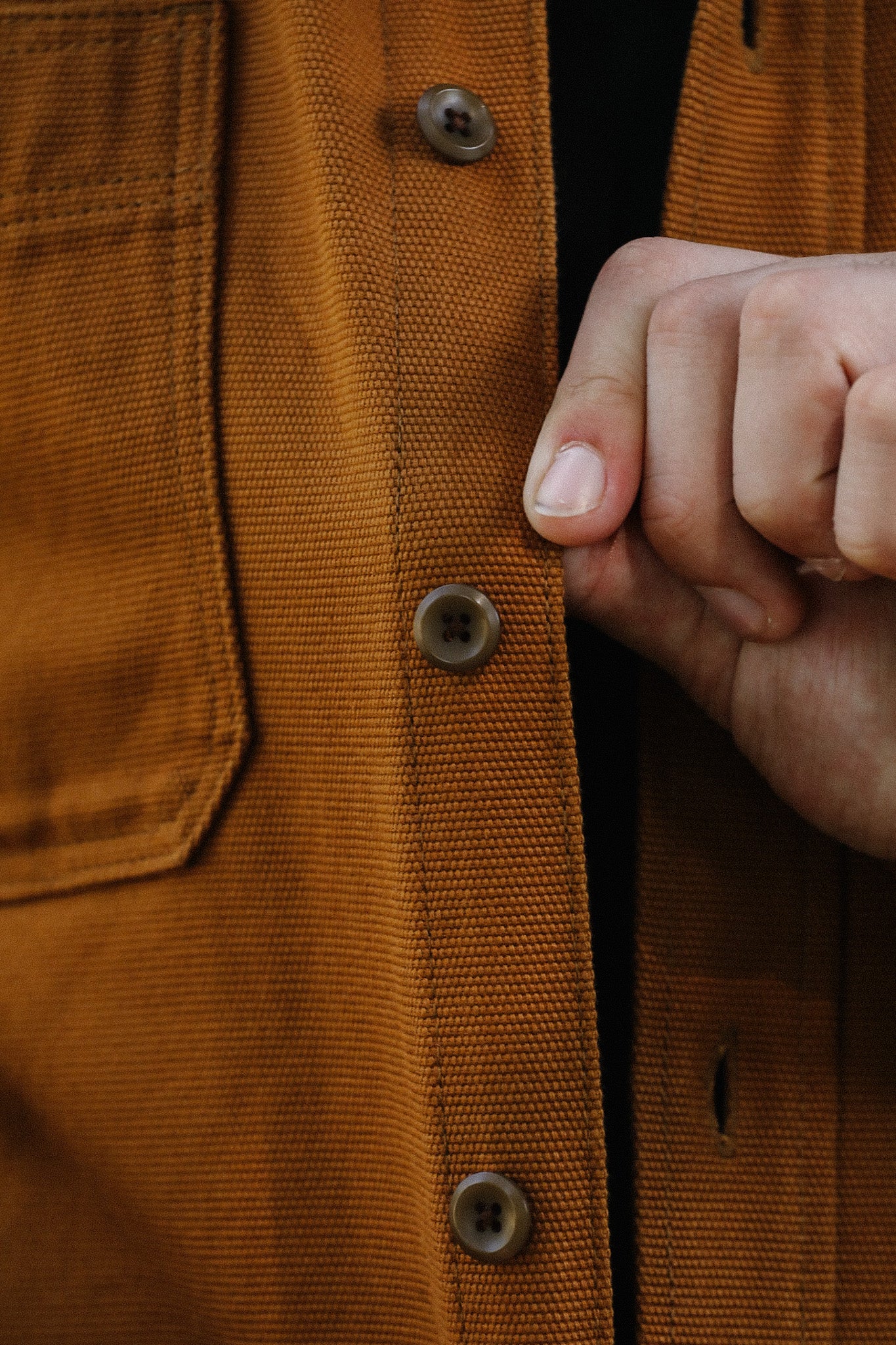 Close-up of hand buttoning a brown textured work shirt with a front pocket, showcasing durable fabric