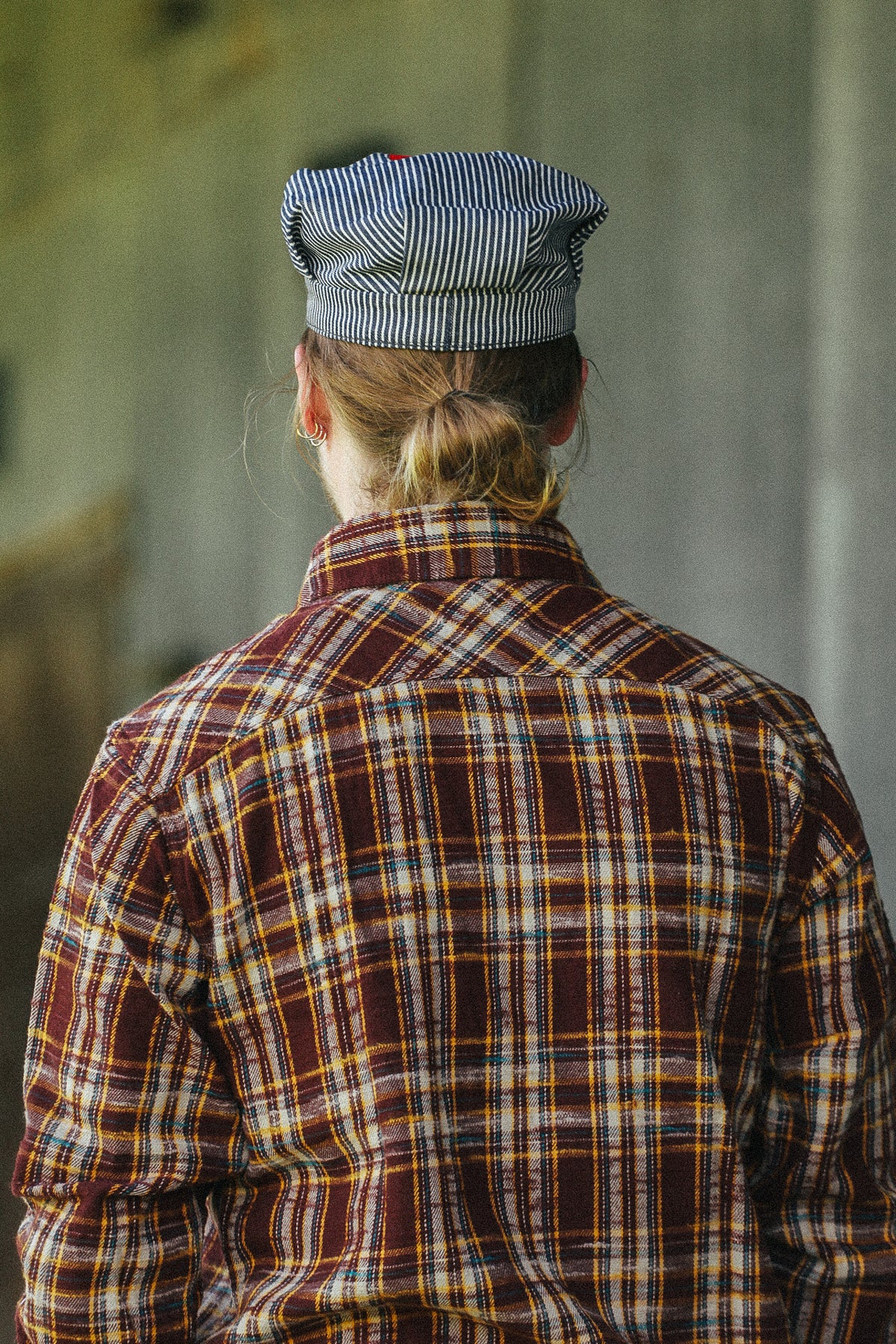 A person viewed from behind wearing a brown and red plaid flannel shirt with yellow and white stripes, and a blue-and-white striped cap, standing against a blurred gray background.