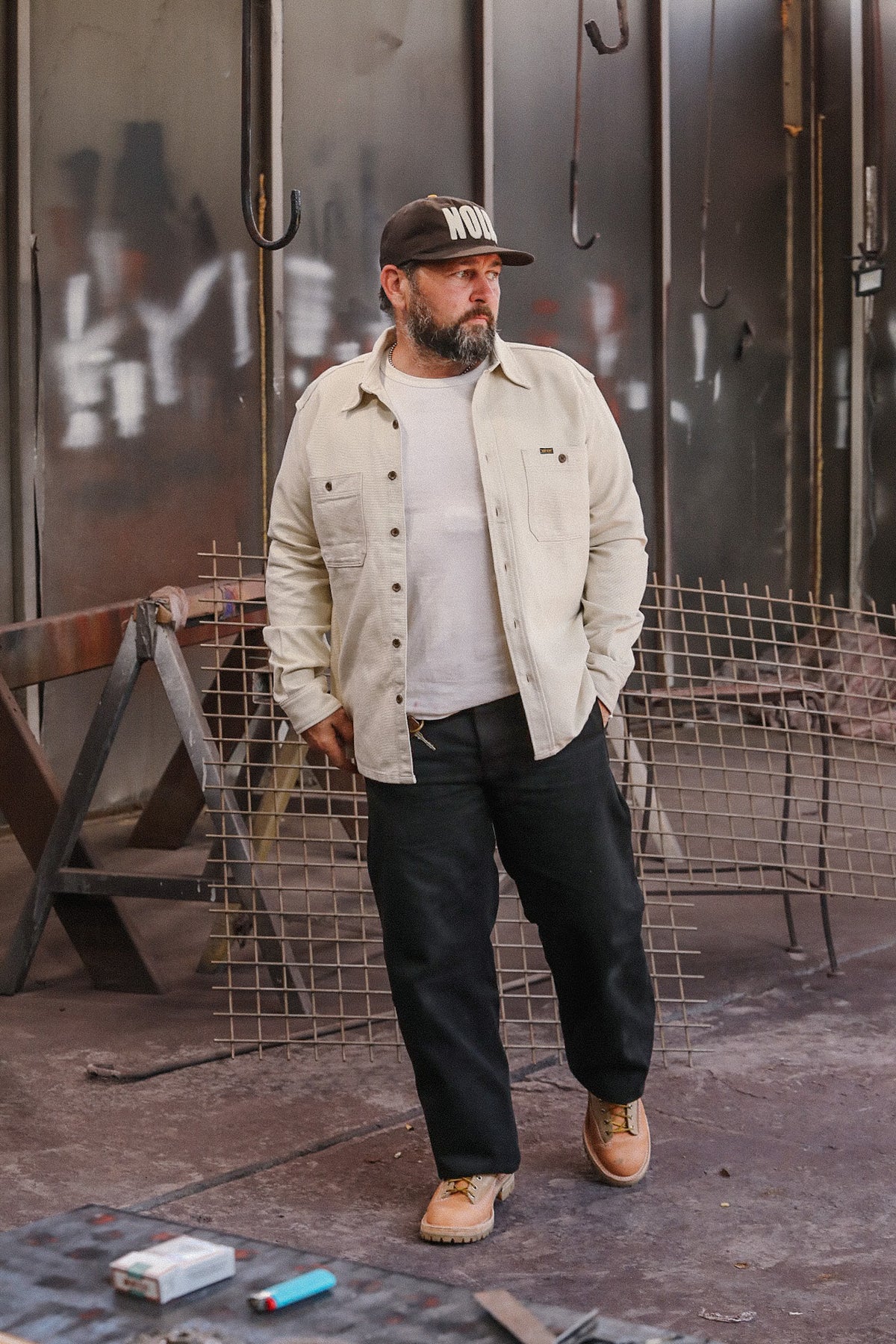Man wearing cream work shirt and black pants in industrial workshop with metal mesh and hooks
