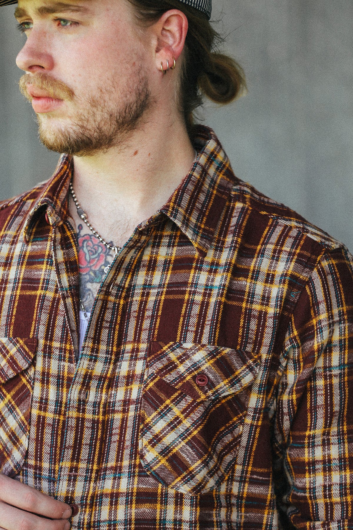 A close-up shot of a man wearing a brown, yellow, and white plaid flannel shirt, looking off to the side against a gray concrete wall.