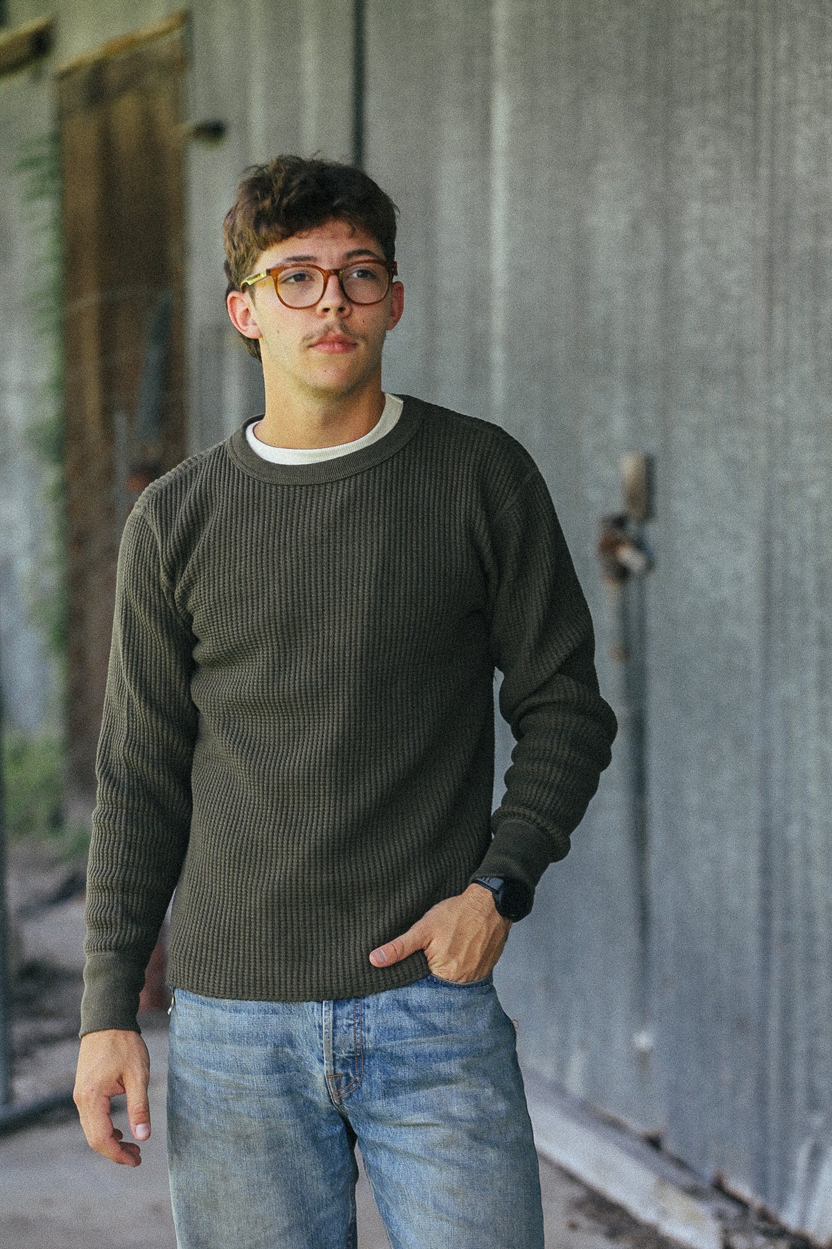 A young man wearing a dark olive green ribbed knit sweater over a white undershirt and blue jeans, standing in front of a weathered gray metal wall.