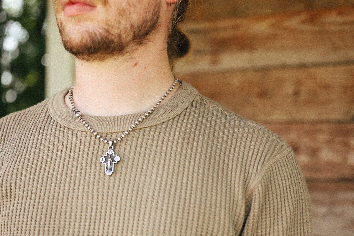 A close-up of a man wearing a silver beaded chain necklace with a silver cross pendant, styled with a beige ribbed knit shirt against a rustic wooden wall background.