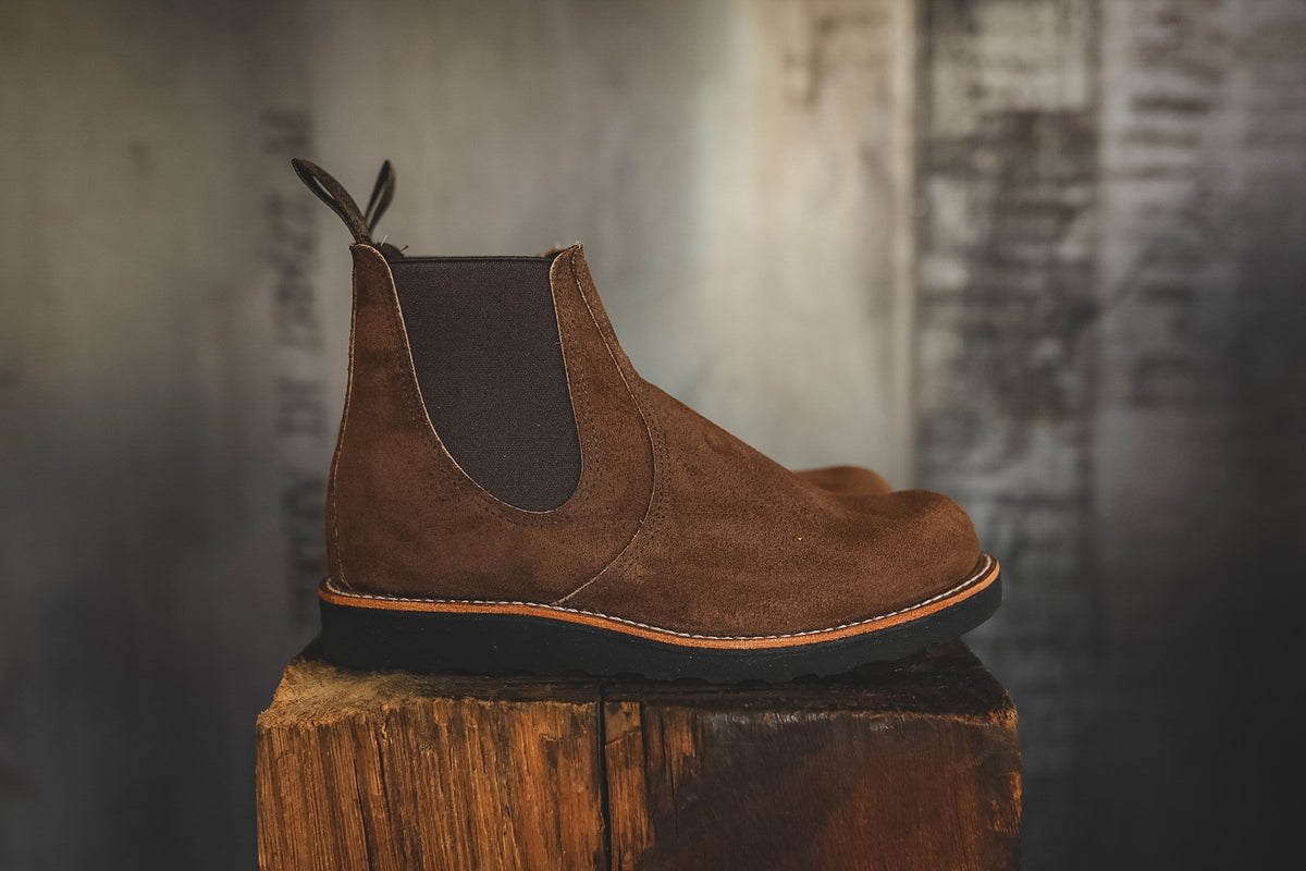 A brown suede Chelsea boot with dark elastic side panels, displayed in profile on a rustic wooden block against a blurred gray background.
