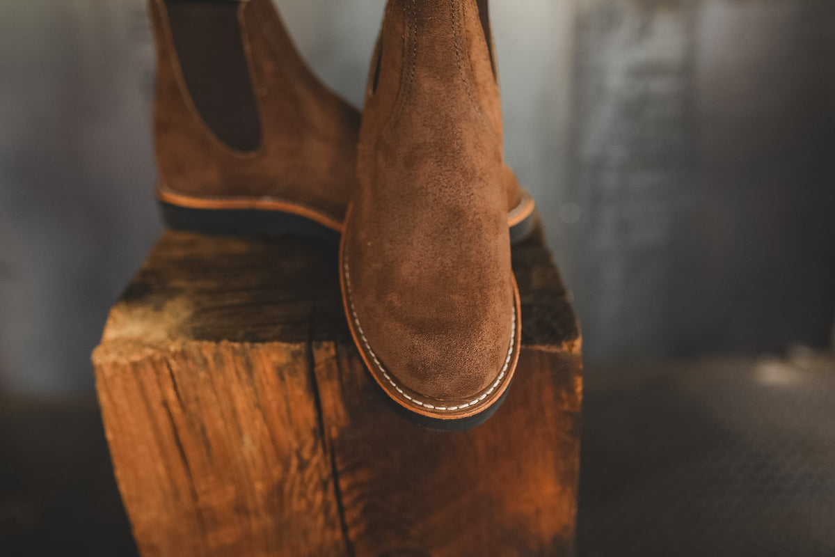 A pair of brown suede Chelsea boots with dark elastic side panels, displayed on a rustic wooden block against a blurred gray background.