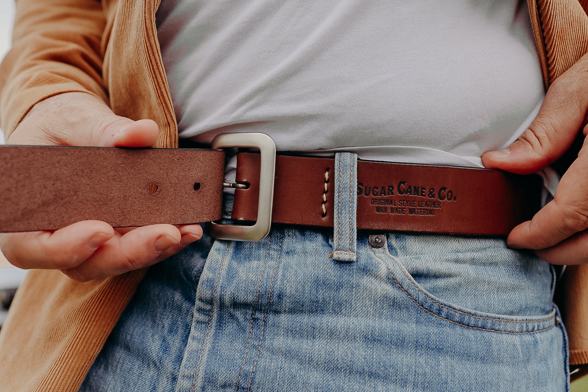 Close-up of brown leather belt with Sugar Cane & Co. logo worn with blue jeans and beige jacket