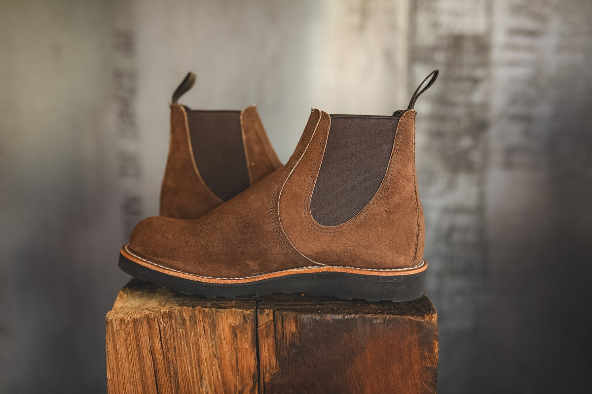 A pair of brown suede Chelsea boots with dark elastic side panels, displayed on a rustic wooden block against a blurred gray background.