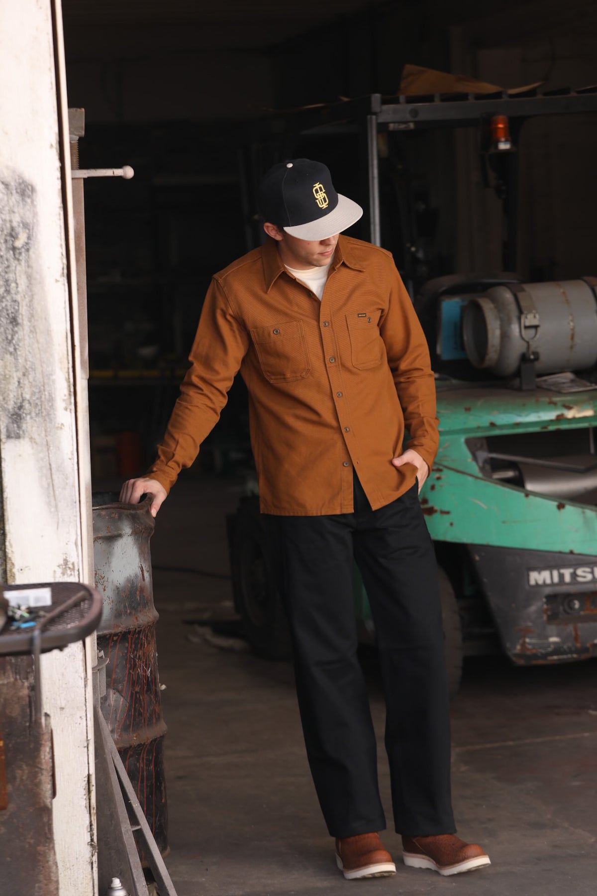 Man wearing brown work shirt, black pants, and baseball cap standing in industrial warehouse with forklift