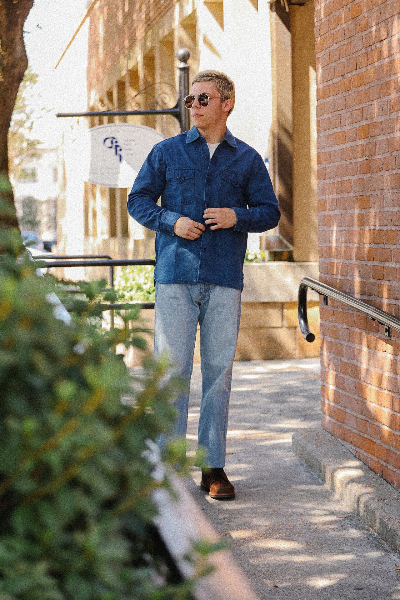 Man wearing blue jacket, sunglasses, light jeans, and brown boots walking along sunny brick sidewalk